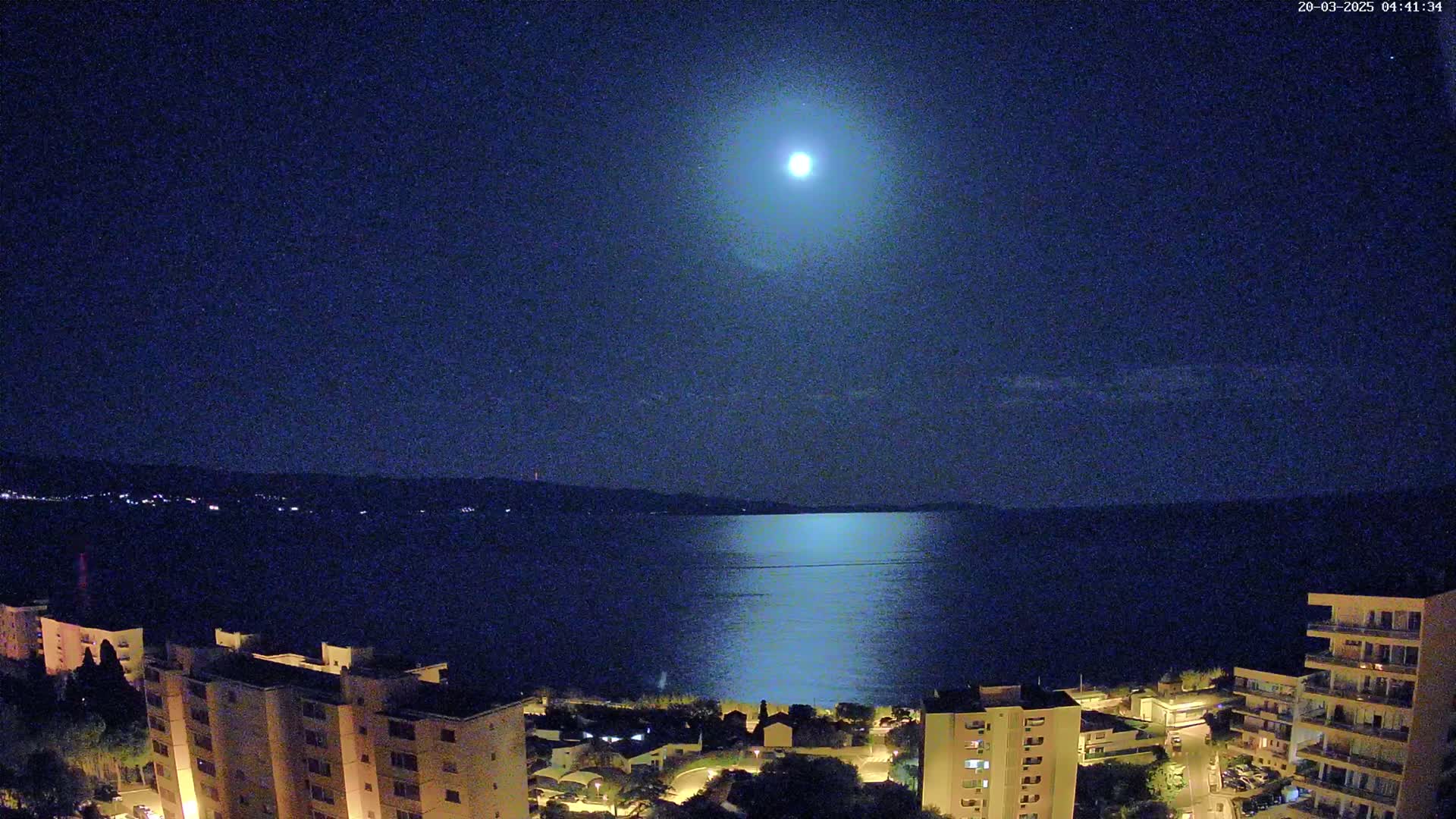 A bright moon illuminates a calm body of water at night, with buildings and a shoreline visible in the foreground.