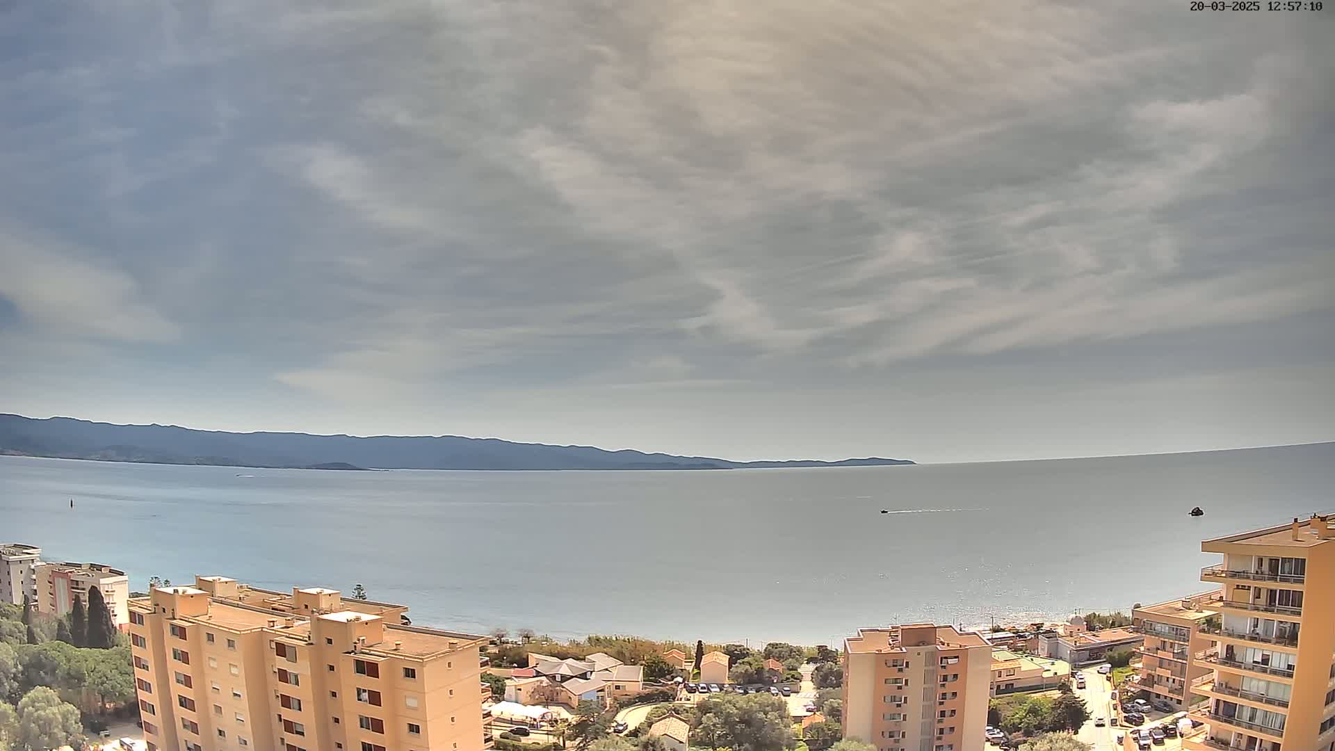 An expansive view of a calm sea under a mostly cloudy sky shows a shoreline with several multi-story buildings and a distant mountain range.