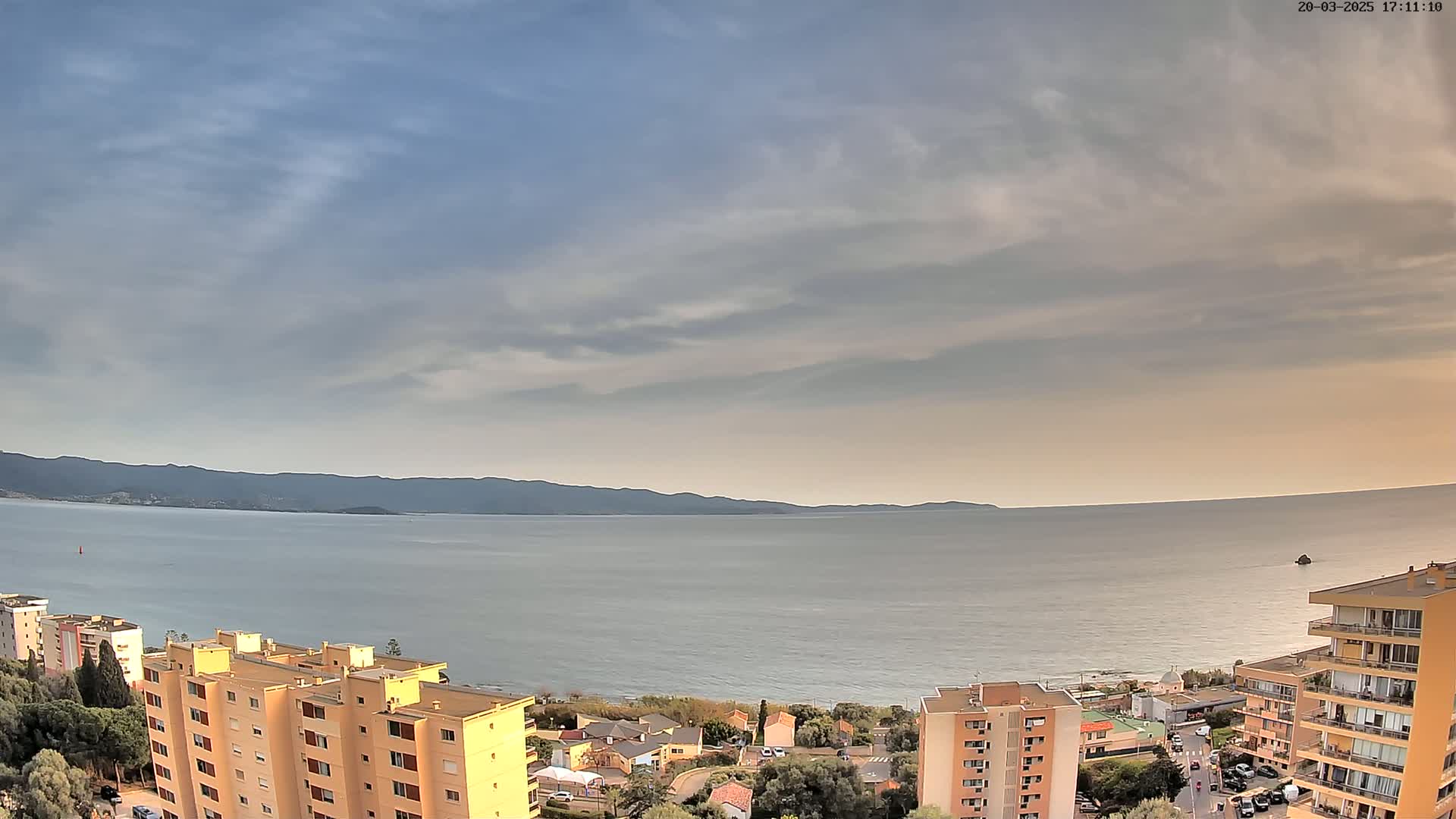 A partly cloudy sky overlooks a calm sea, with a coastal town and distant mountains visible.