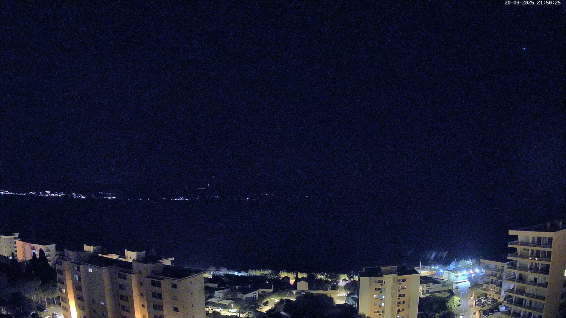 A nighttime view shows a dark body of water with a distant, faintly lit shoreline, overlooked by several multi-story buildings.