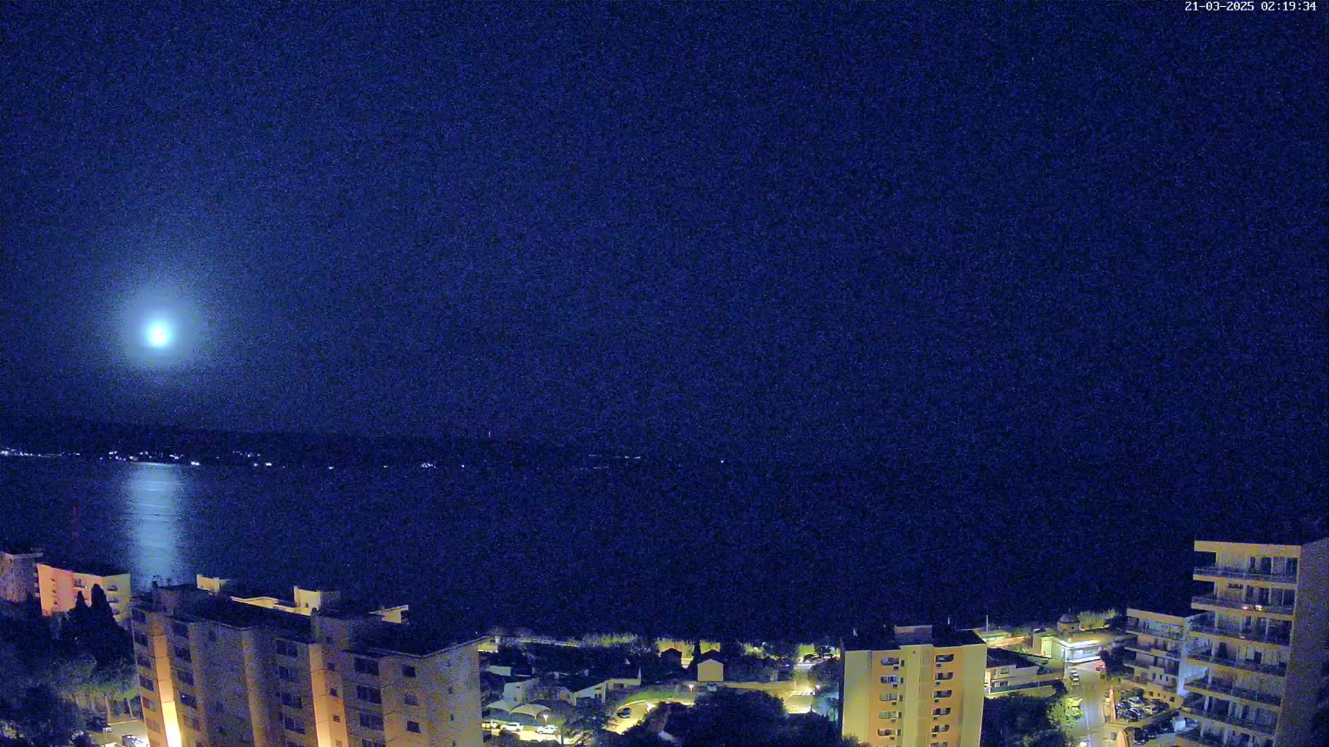 A bright moon illuminates a calm body of water at night, with buildings and lights visible along the shoreline.