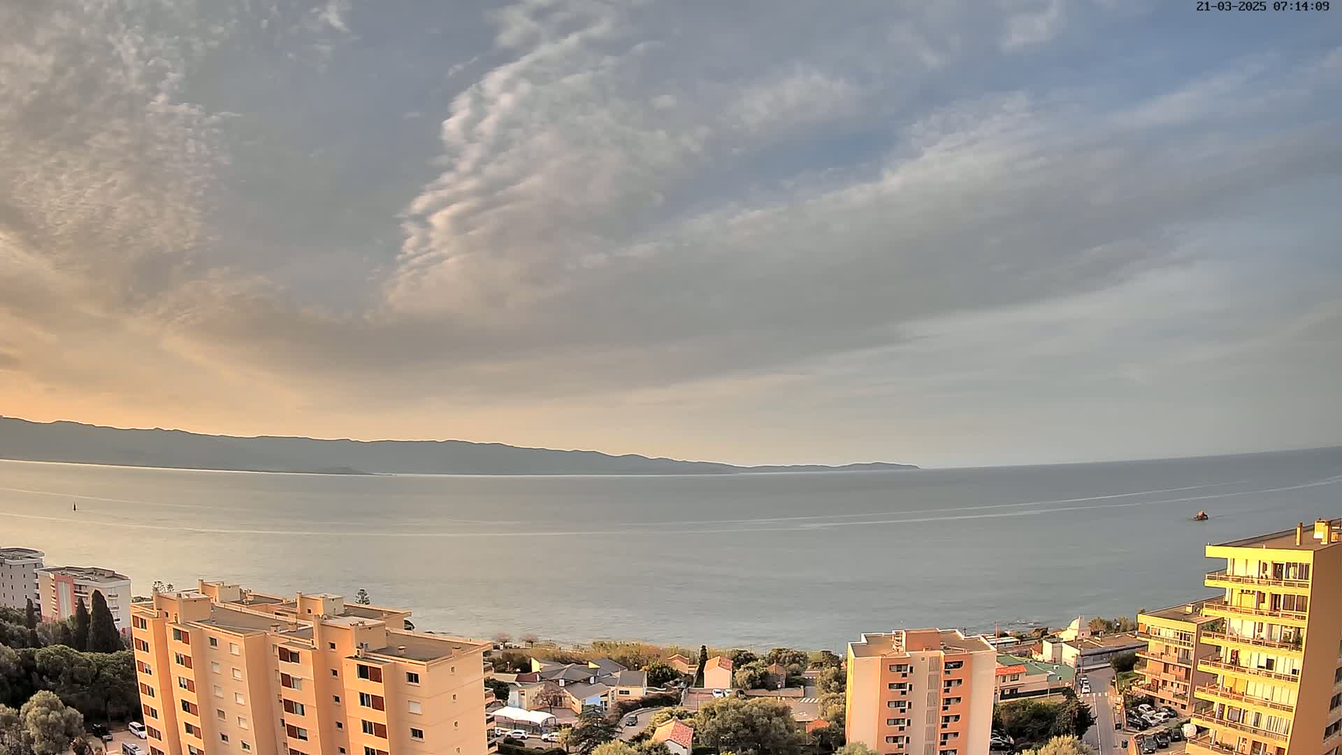 An elevated view shows a calm sea under a partly cloudy sky, with apartment buildings and a shoreline visible in the foreground.