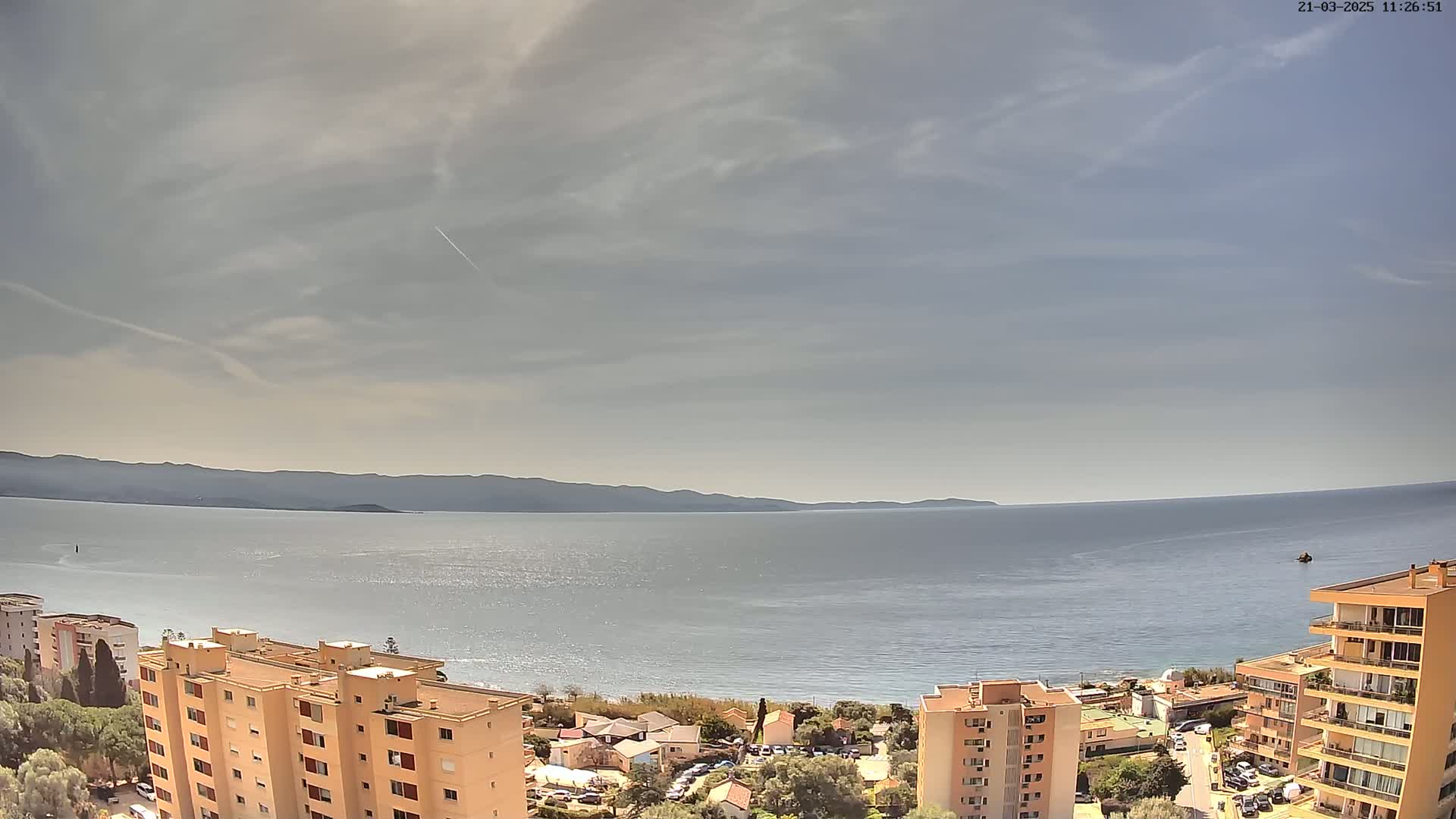 A mostly sunny day overlooking a calm sea with distant mountains and several apartment buildings in the foreground.