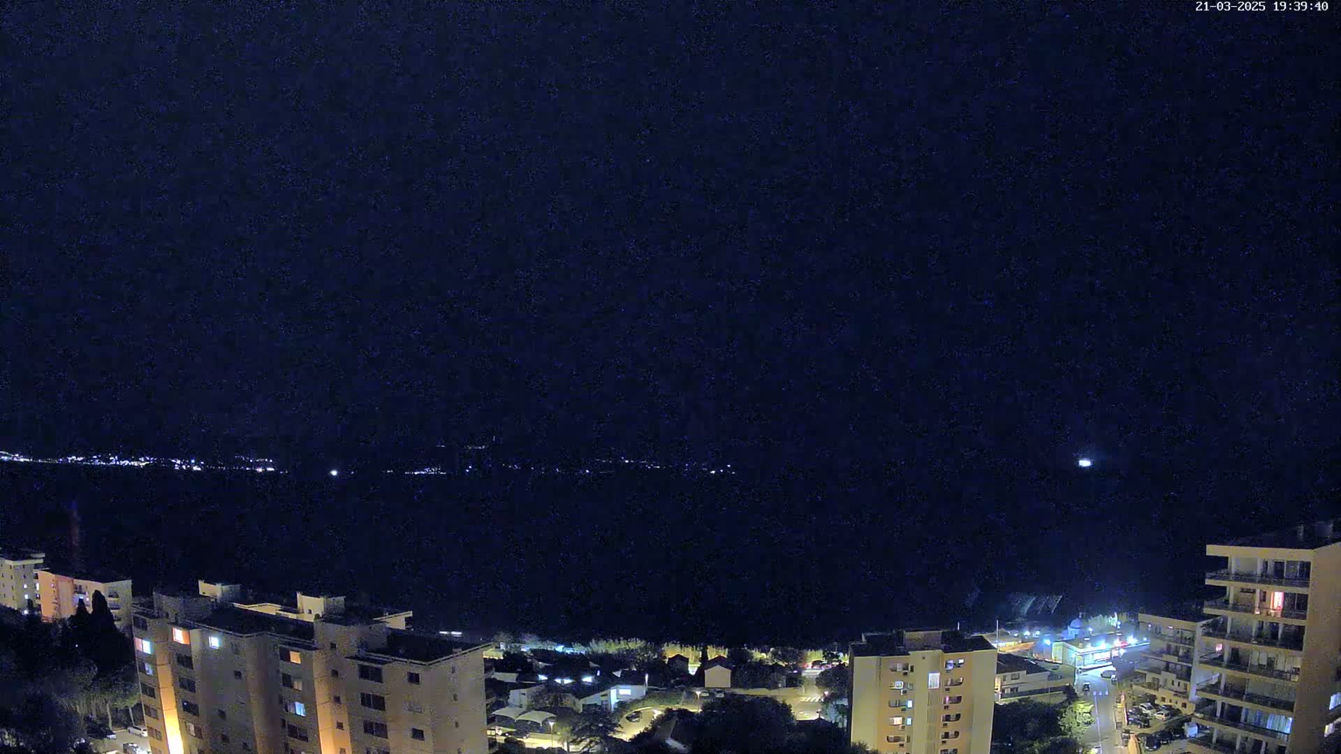 A nighttime view shows a dark body of water with a distant, faintly lit shoreline, overlooked by several illuminated buildings under a clear, star-studded sky.
