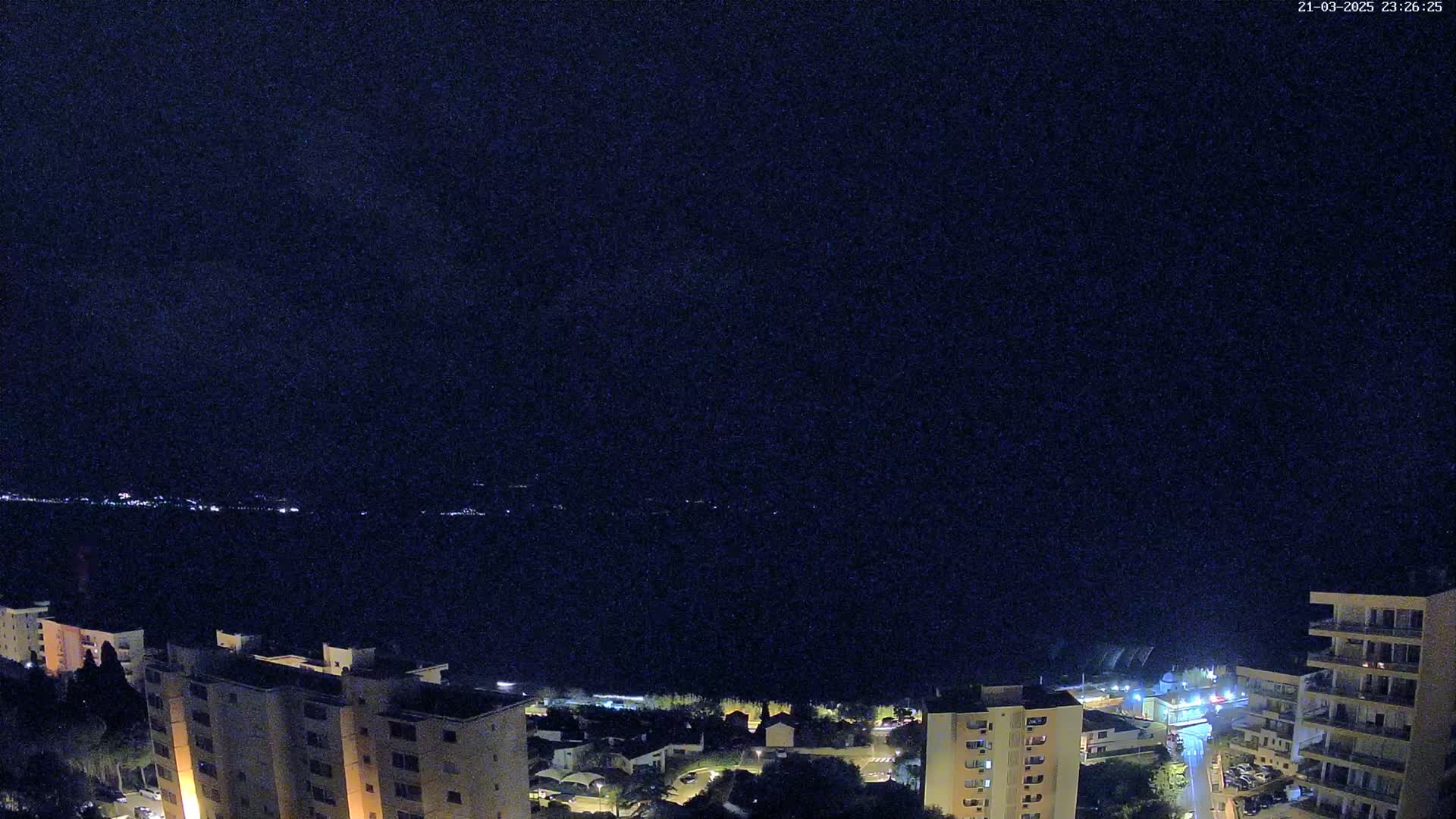 A nighttime view of a dark body of water, with a town's illuminated buildings and streets visible along the shore under a clear, dark sky.