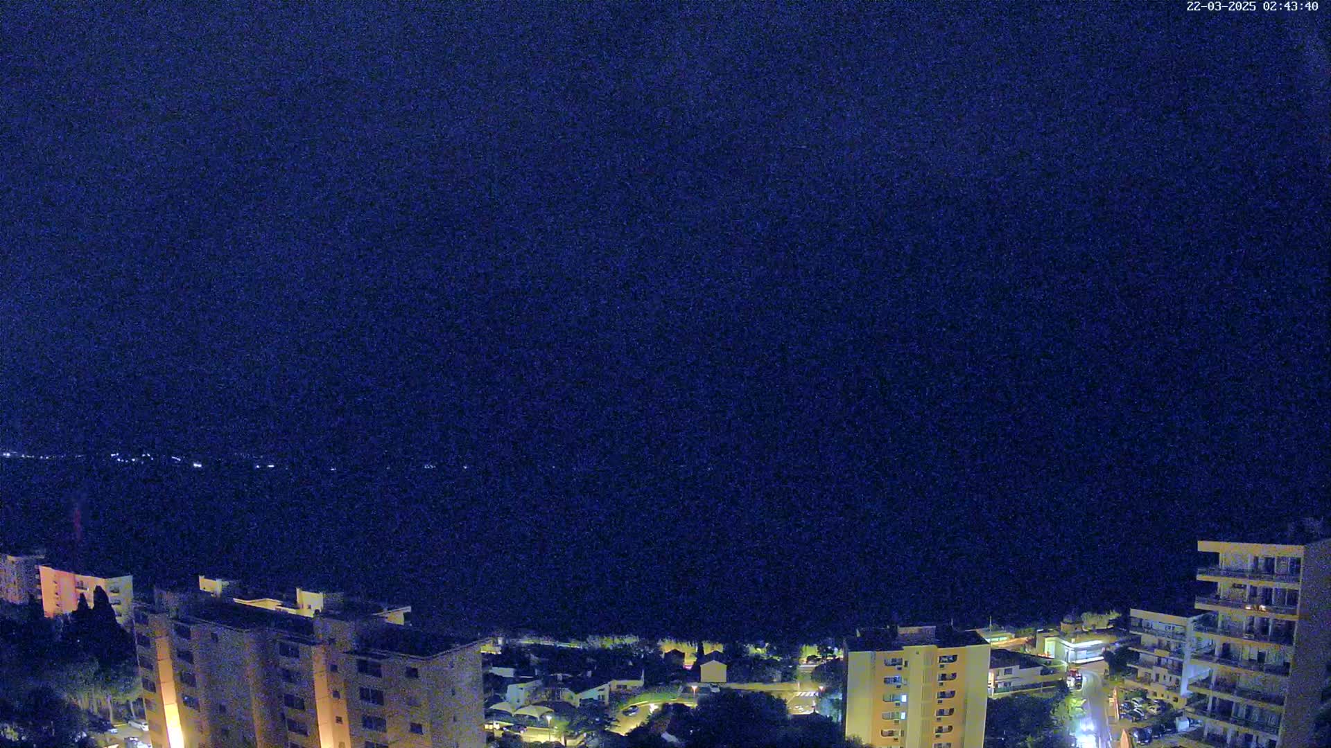 A nighttime aerial view shows a coastal town with several apartment buildings under a dark, clear sky.