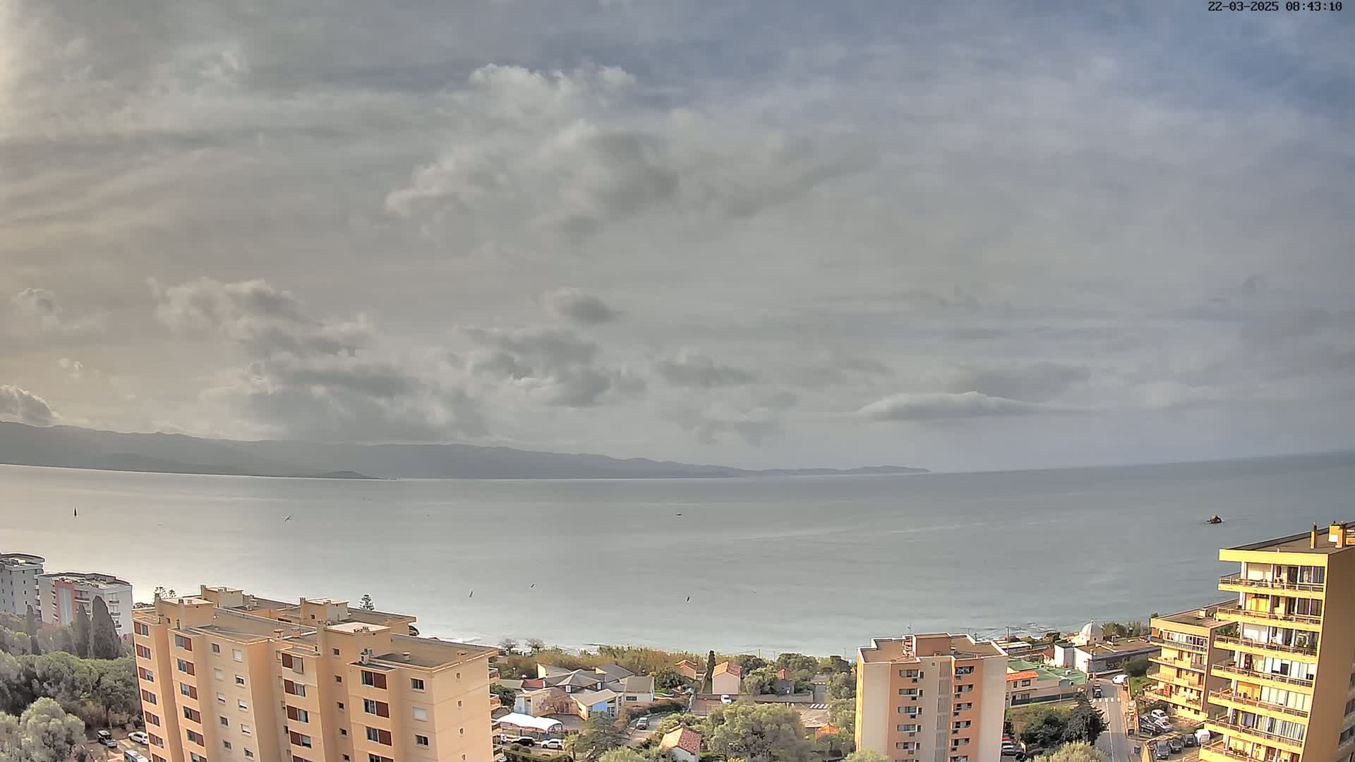 An overcast sky hangs over a calm sea, with a coastal town and several apartment buildings nestled between the water and distant mountains.
