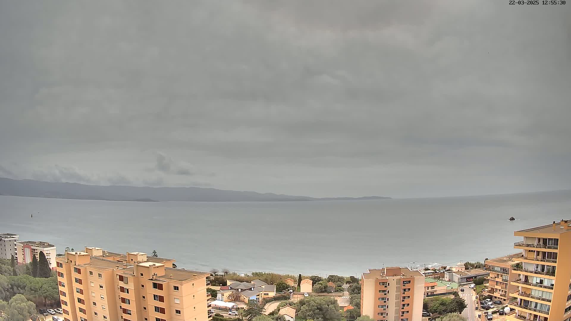 An overcast sky hangs over a calm sea, with multi-story buildings and a coastal town visible in the foreground.