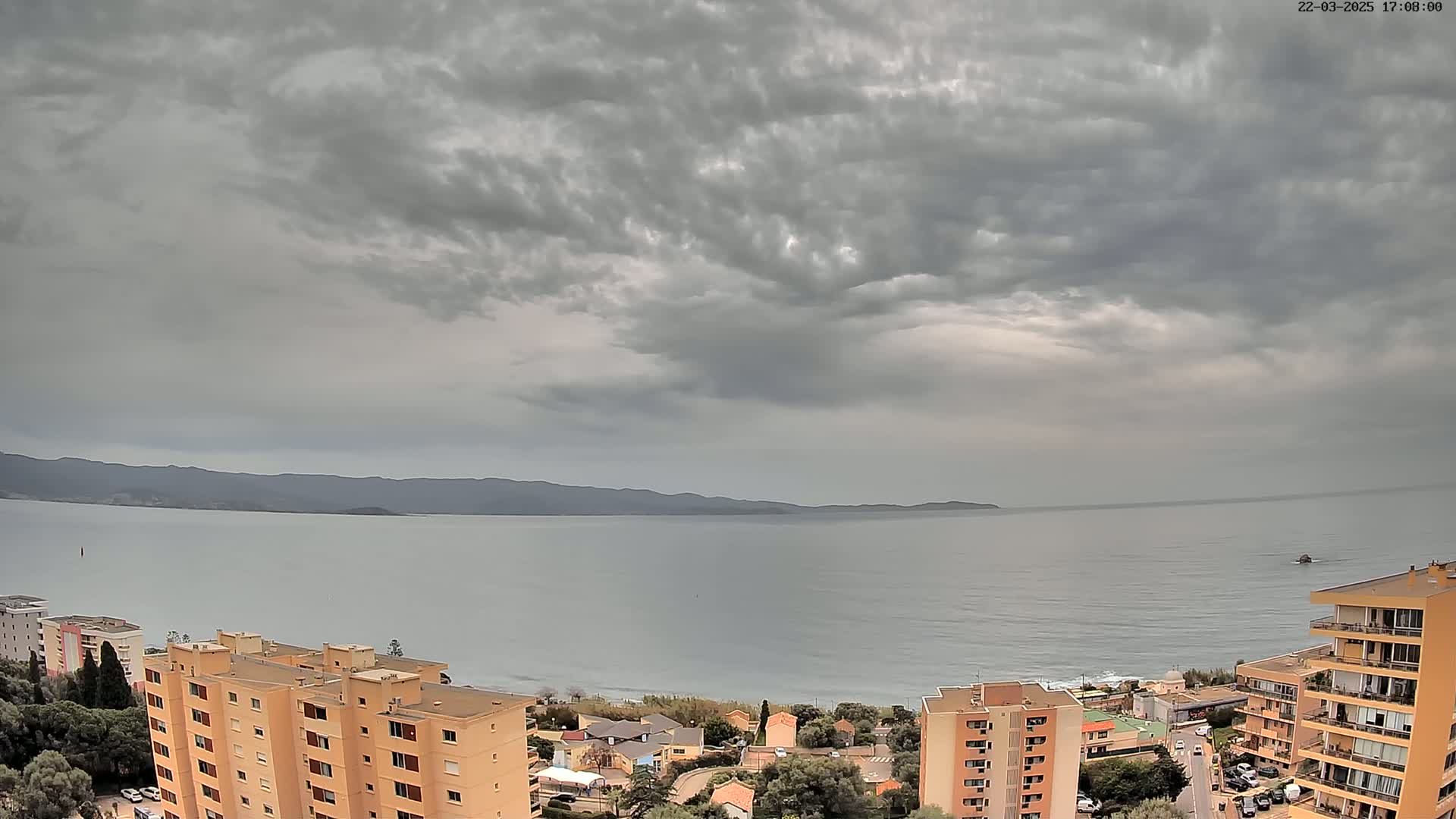 An overcast sky hangs over a calm sea, overlooking a coastal town with several multi-story buildings.