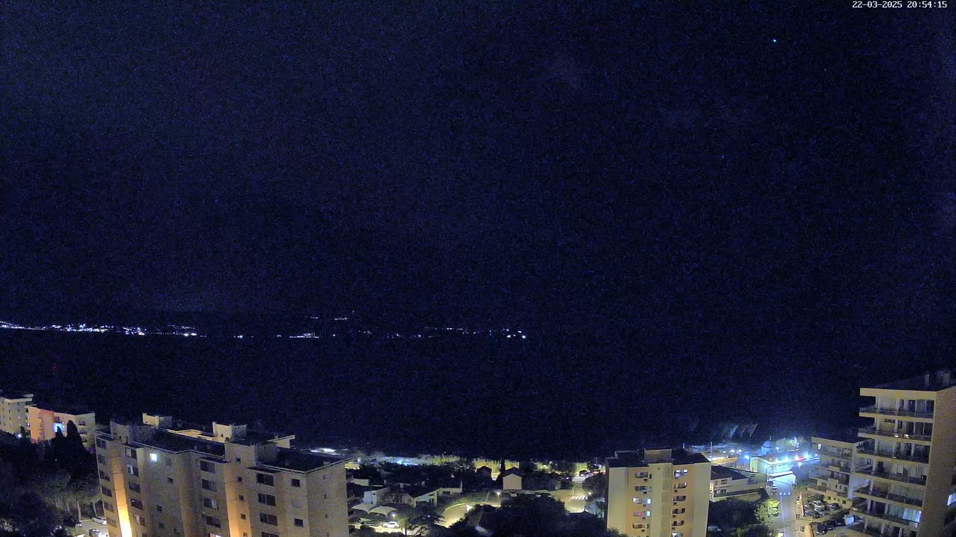 A nighttime, high-angle view shows a dark body of water with a distant, faintly lit shoreline, overlooked by several multi-story buildings illuminated by lights.