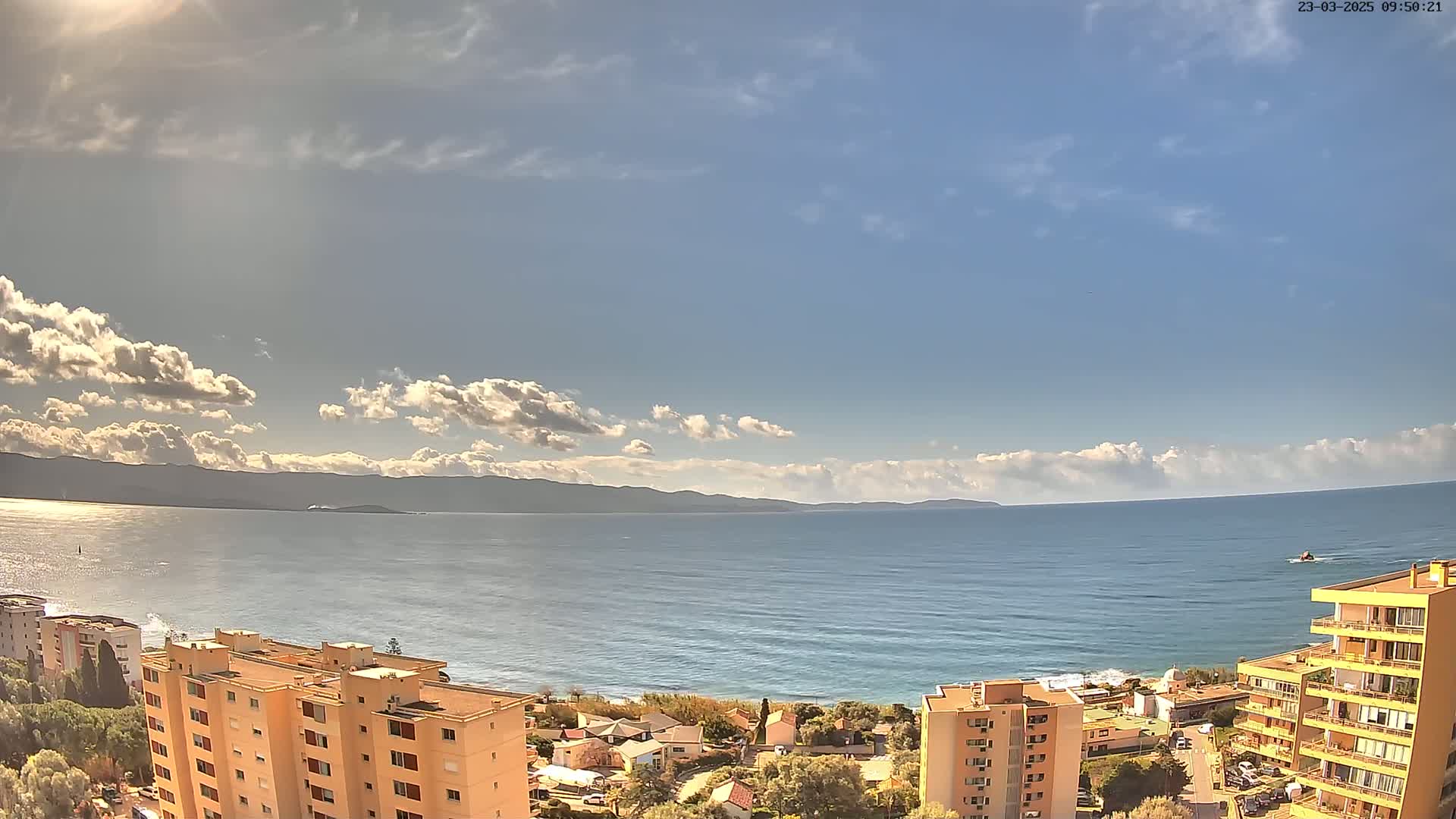 A partly sunny day reveals a coastal scene with calm ocean waters, distant mountains, and several multi-story buildings nestled along the shore.