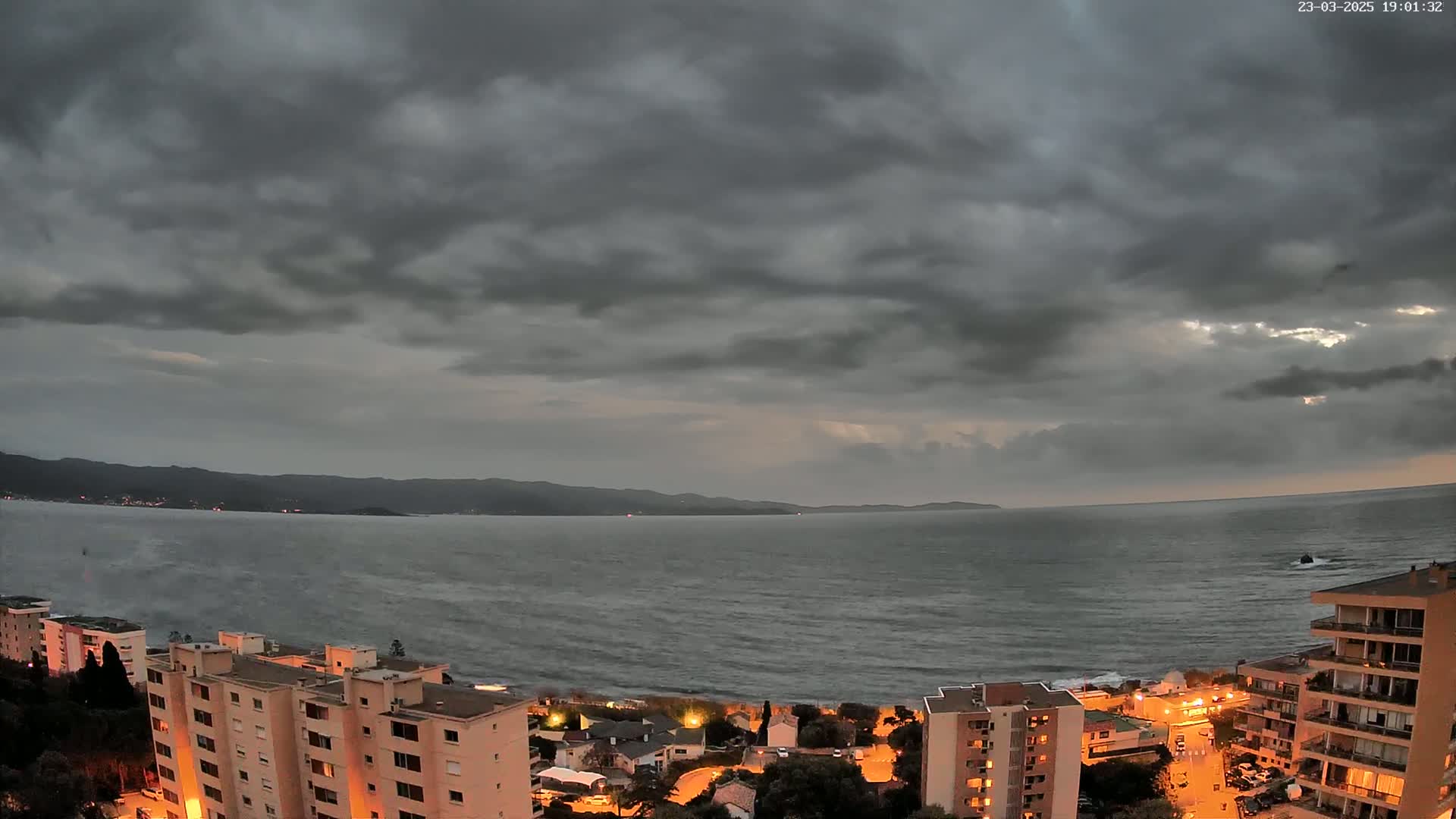 A coastal town at dusk under a dark, cloudy sky overlooks a choppy sea and distant mountains.