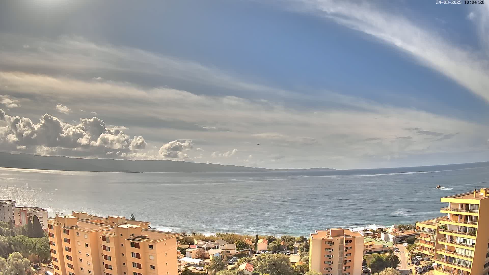 A partly cloudy day overlooks a calm sea with a distant mountain range and coastal buildings.