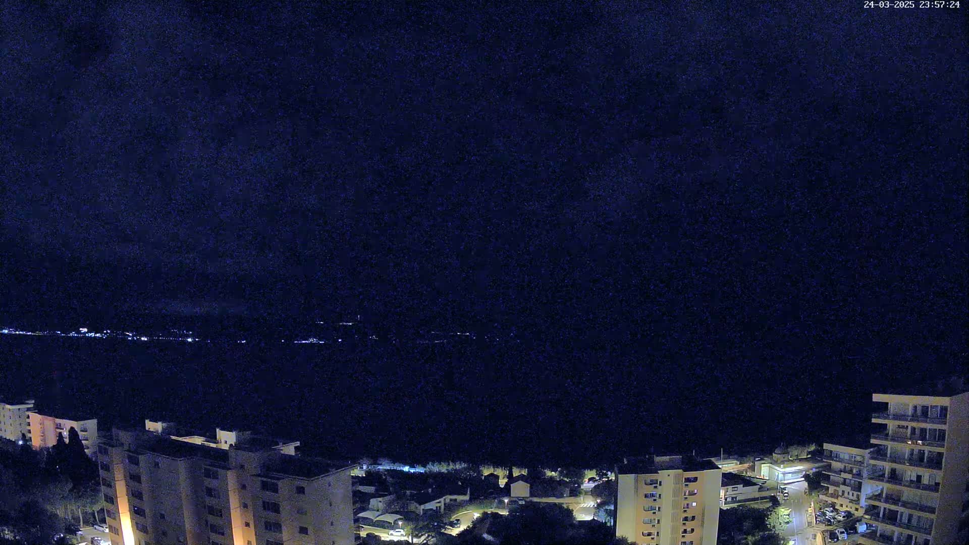 A nighttime view of a coastal town with several apartment buildings, illuminated by streetlights, overlooking a dark body of water under a clear night sky.