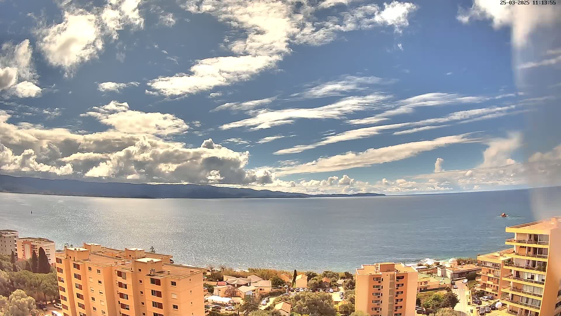 A partly cloudy sky overlooks a calm sea with a distant mountain range, and several apartment buildings are situated along the shore.