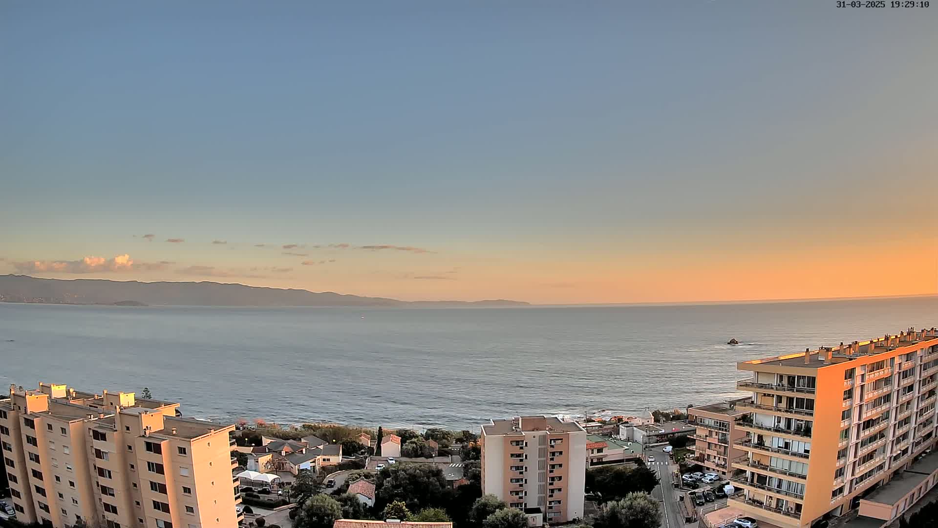 A coastal sunset view shows a calm sea extending to distant mountains under a pale orange and blue sky, with several apartment buildings situated on a hillside overlooking the water.