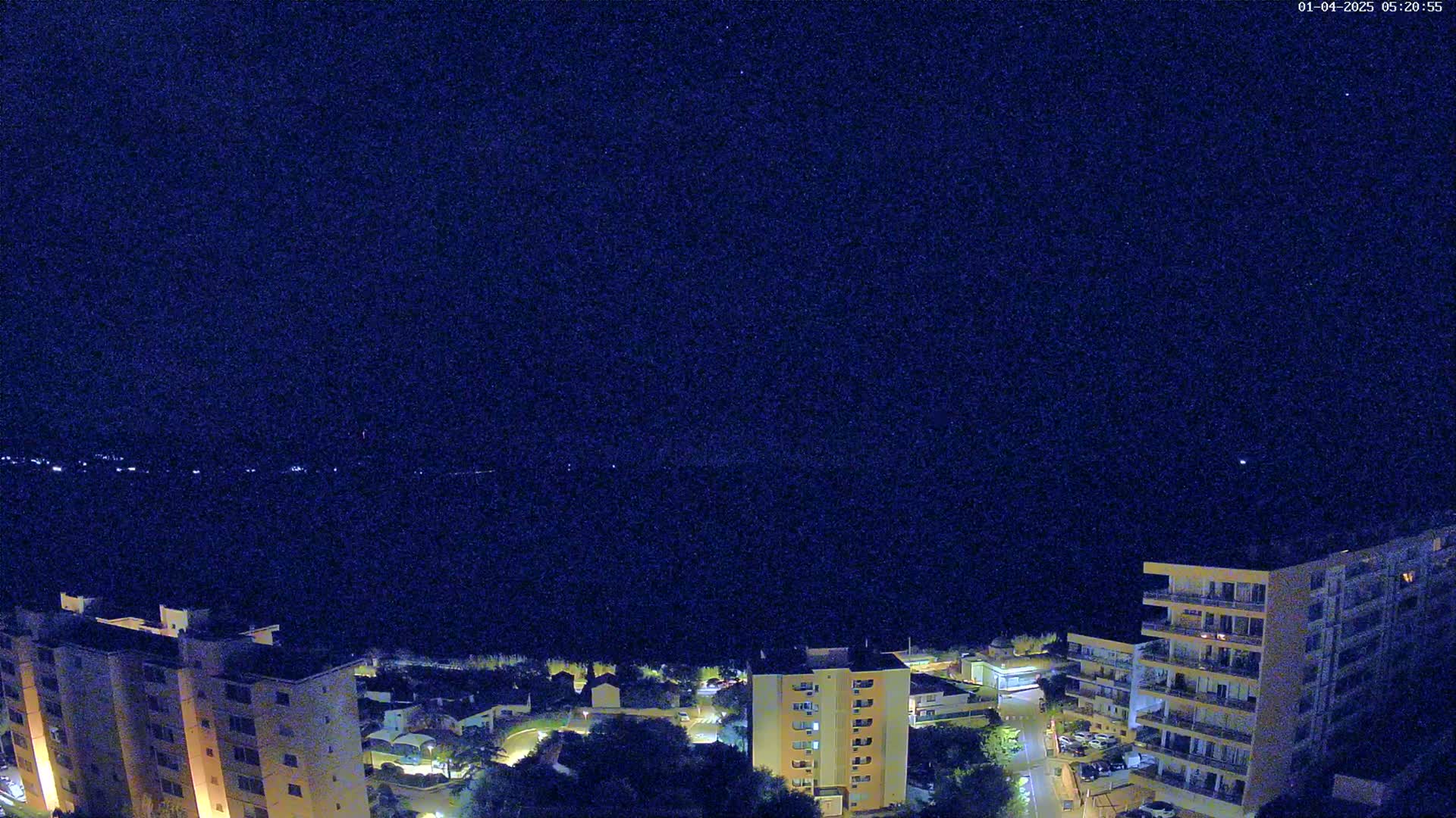 A nighttime, high-angle view shows several multi-story buildings illuminated, overlooking a dark body of water under a clear, starry sky.
