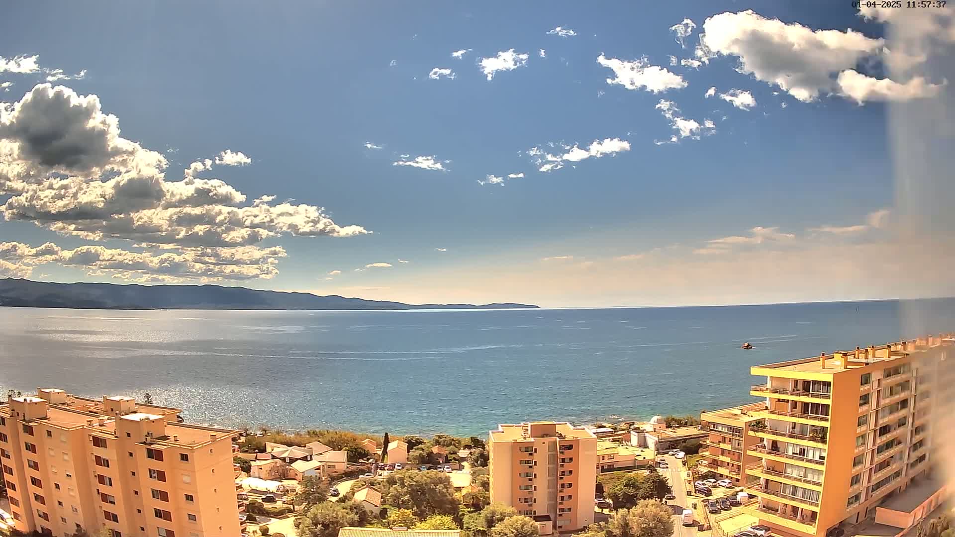 A partly cloudy sunny day overlooks a calm sea with distant mountains, and several buildings are visible in the foreground.