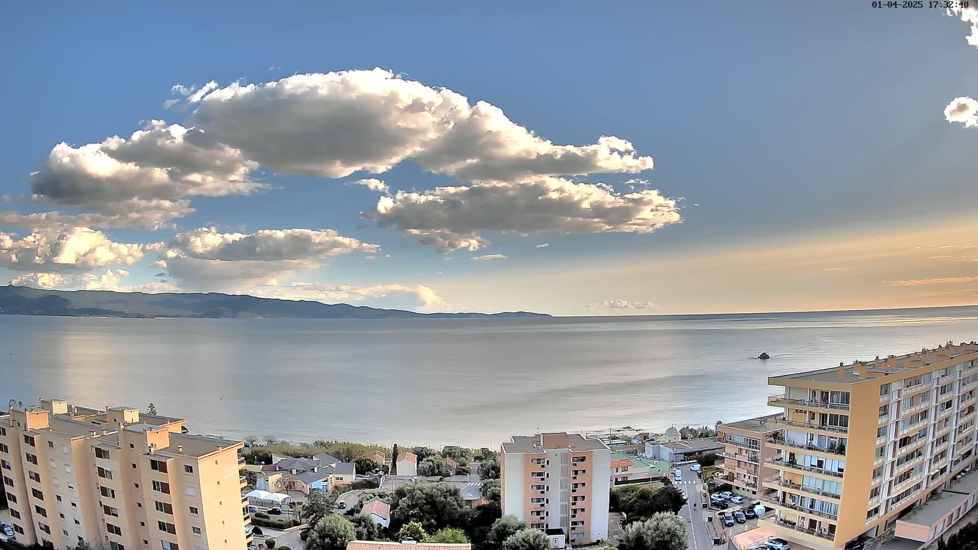 A partly cloudy sky overlooks a calm sea, a beach, and several apartment buildings.