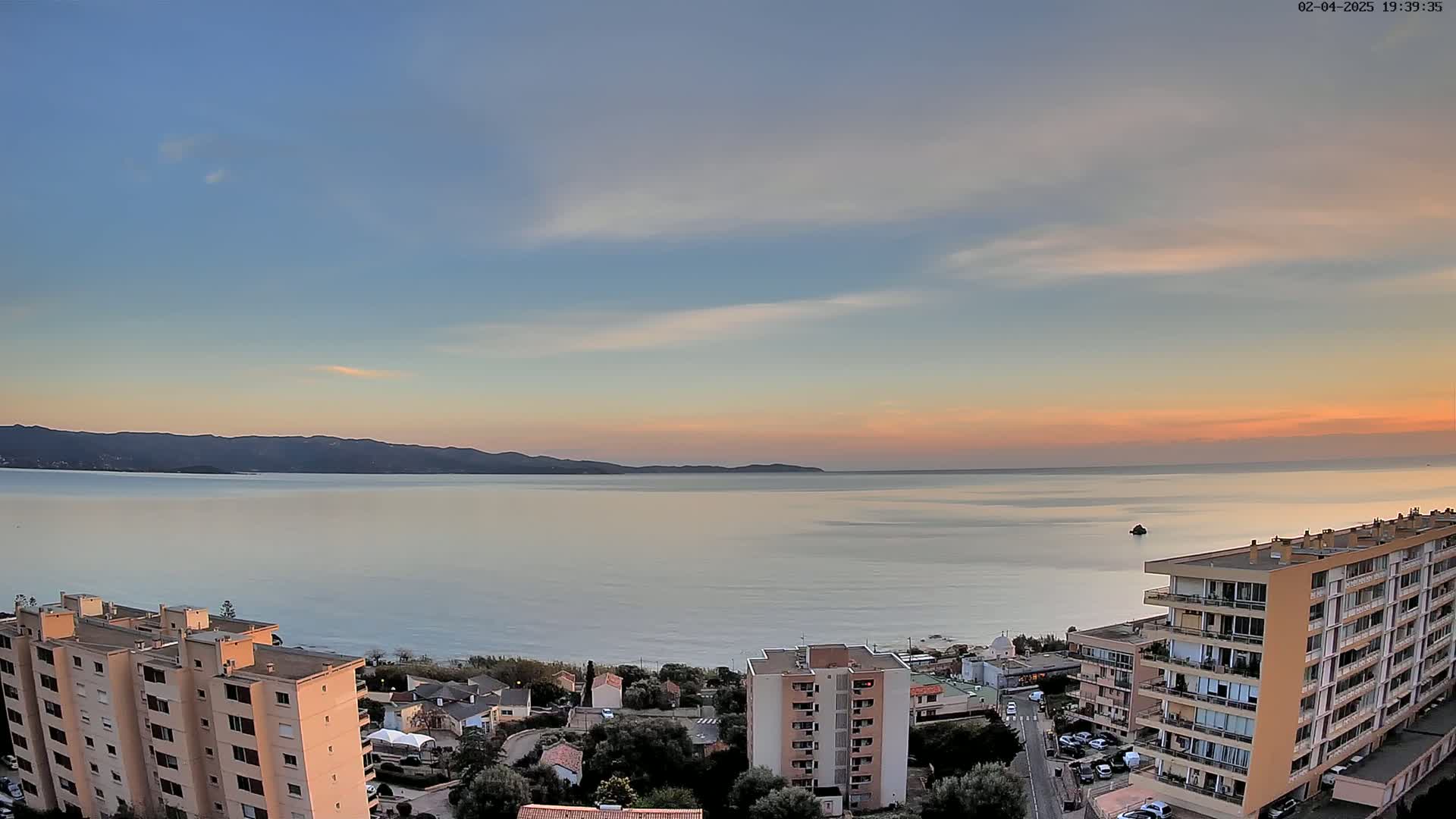 A calm sea at sunset is viewed from above, showing a shoreline with buildings and a distant mountain range under a pale sky.