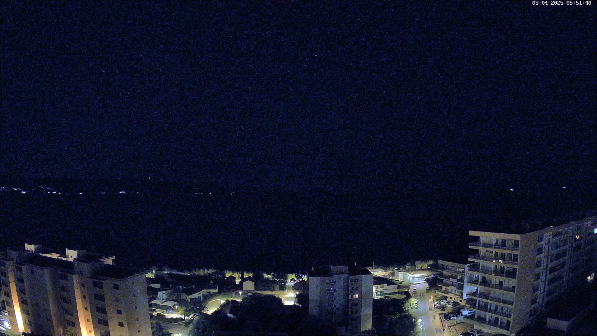 A nighttime view of several illuminated apartment buildings overlooking a dark body of water under a clear, starry sky.