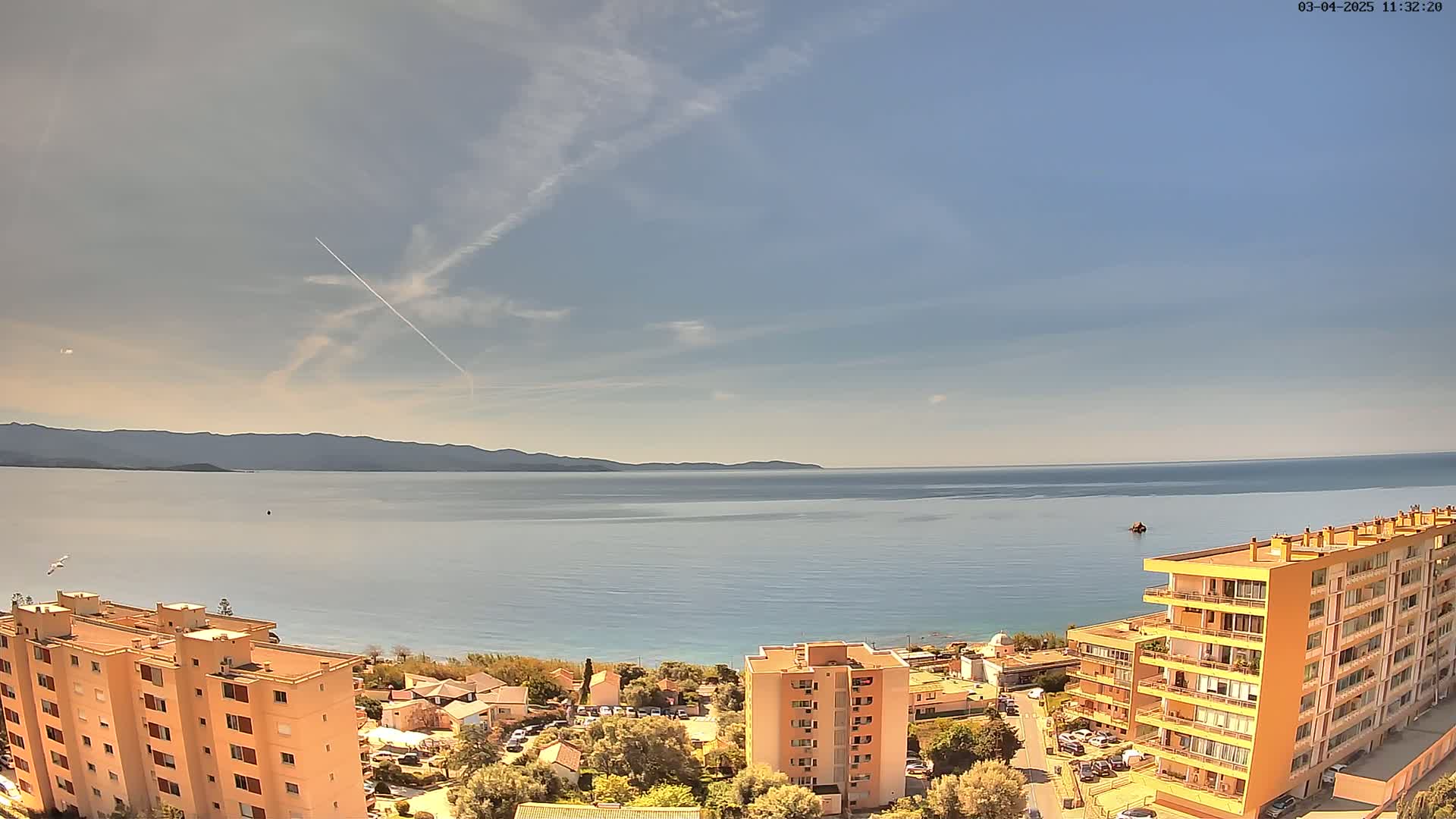An aerial view shows a calm sea under a mostly clear sky with a contrail, overlooking a coastal town with several multi-story buildings.