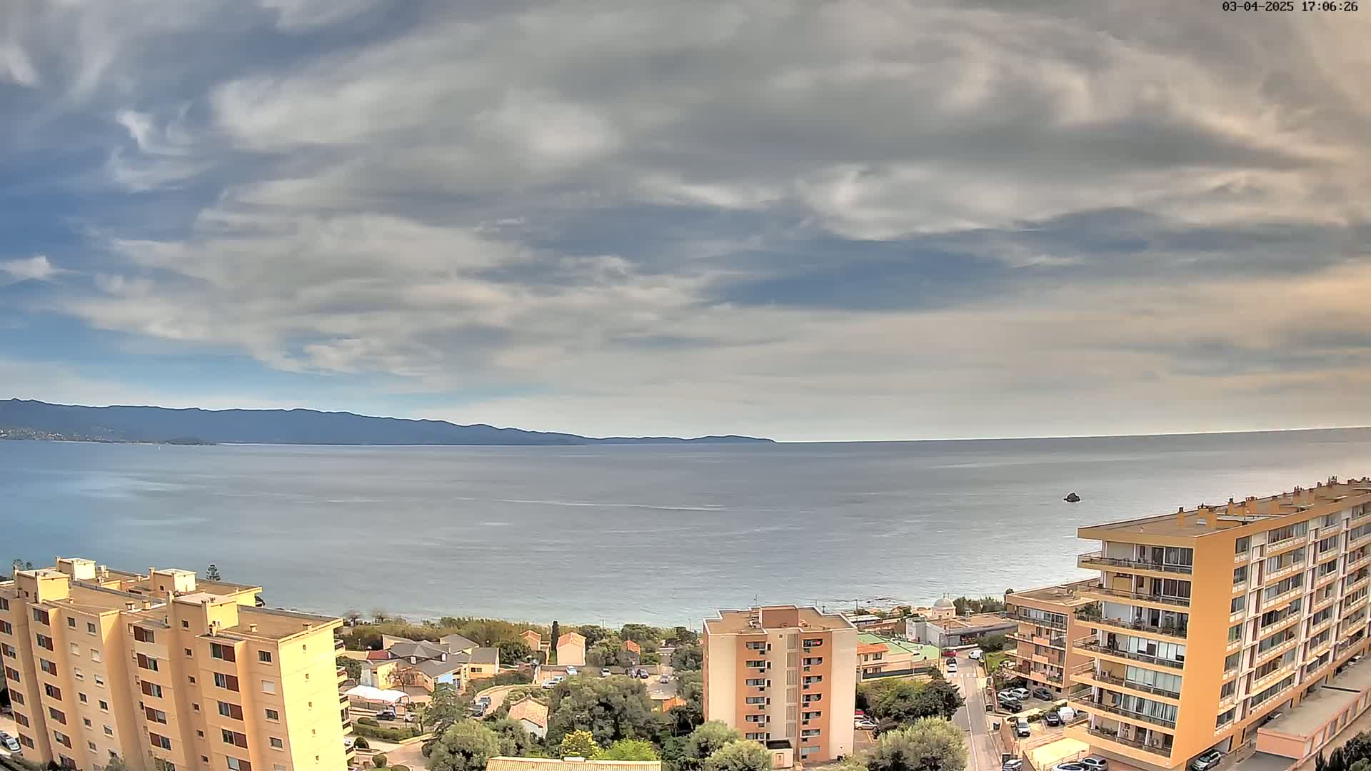 An aerial view shows a calm sea under a partly cloudy sky, with several apartment buildings and some residential areas situated along a coastline backed by distant mountains.