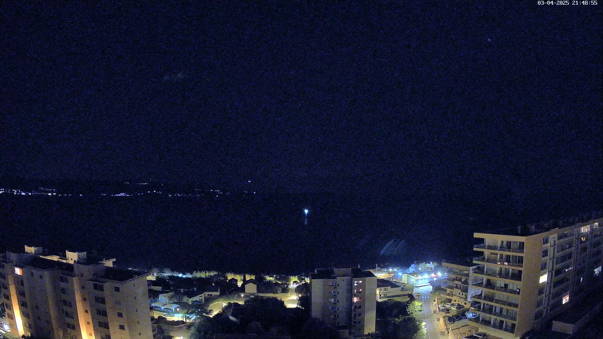 A nighttime view of a coastal town, with several illuminated buildings and a dark body of water extending to a distant, faintly lit shoreline under a clear, starlit sky.