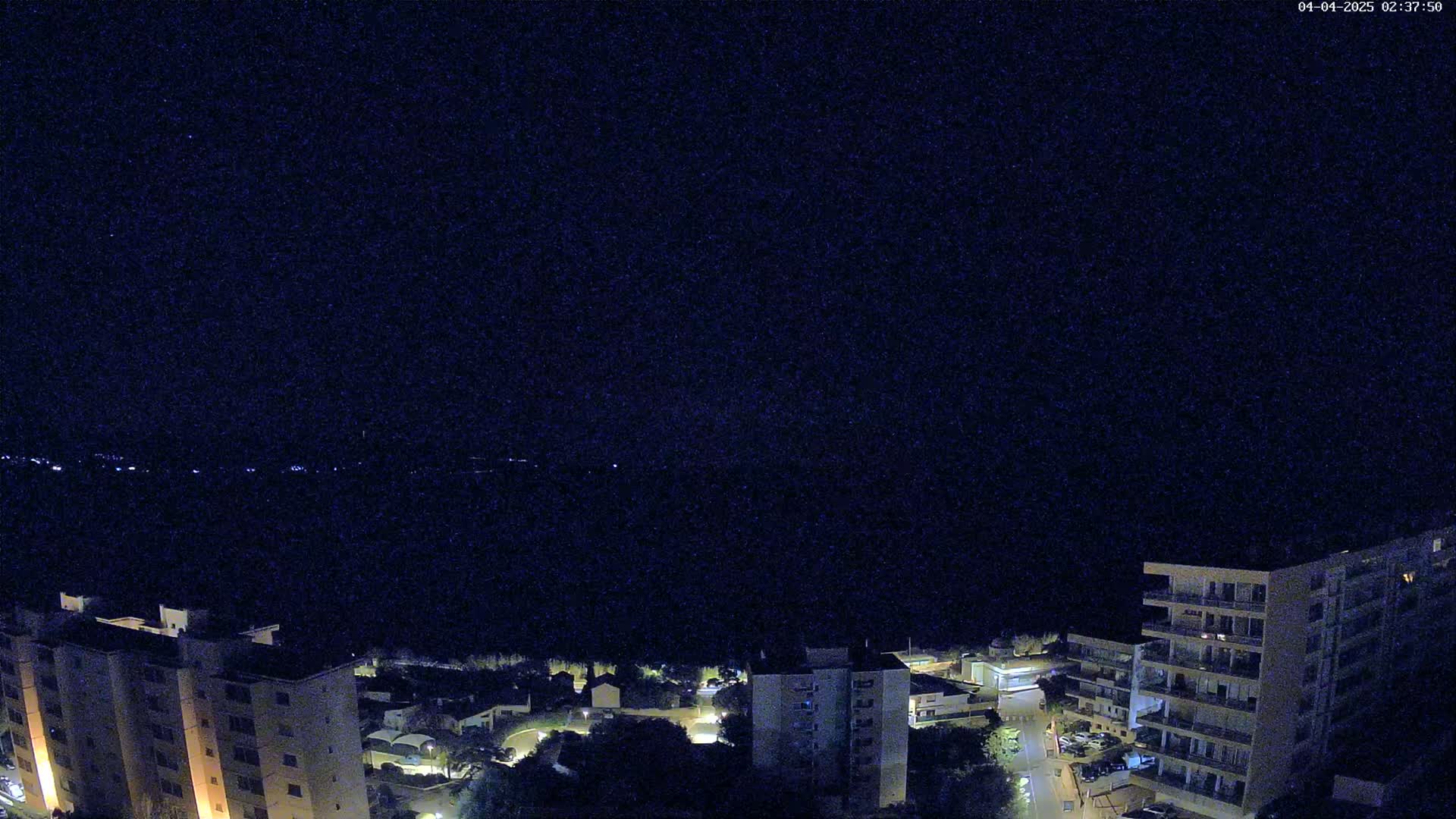 A nighttime aerial view shows several multi-story apartment buildings illuminated against a dark, clear sky over a body of water.