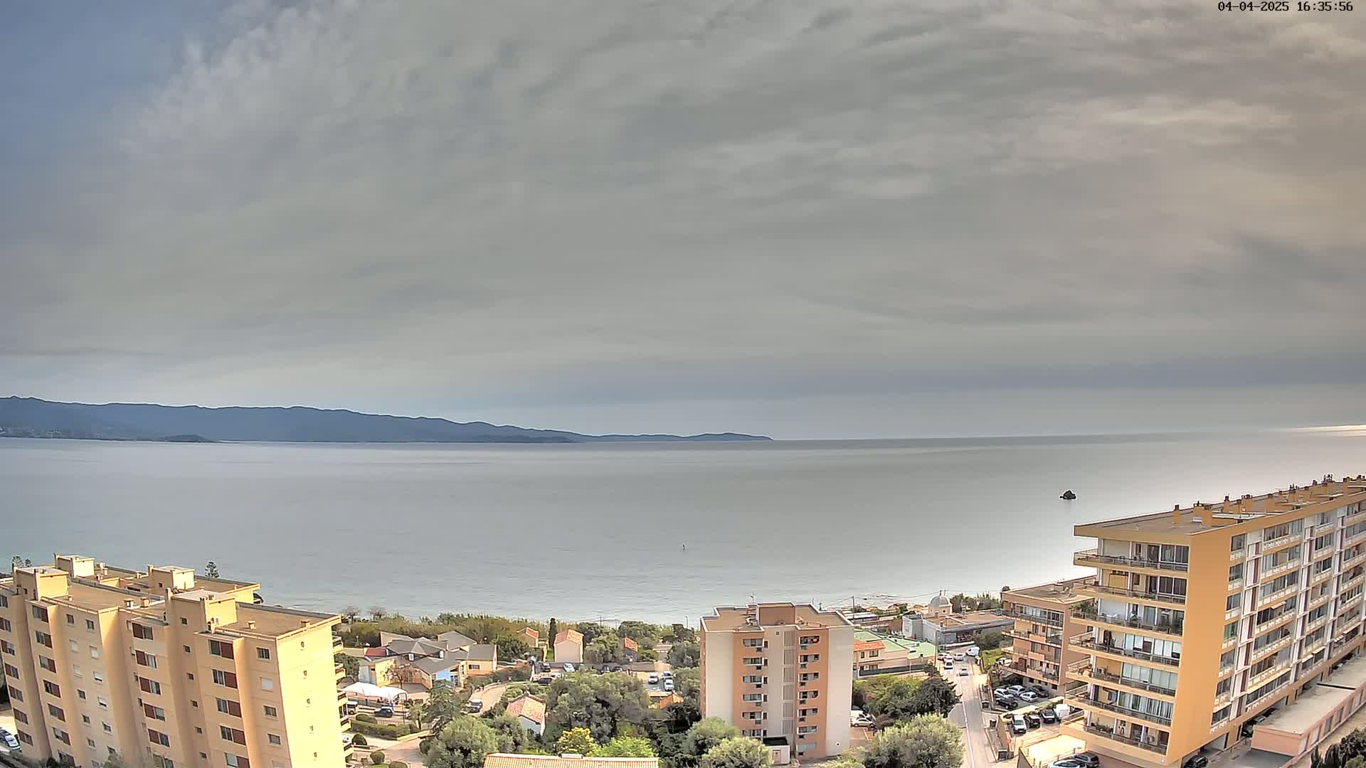 A mostly cloudy day overlooks a calm ocean with a distant mountain range, a sandy beach, and several multi-story buildings.