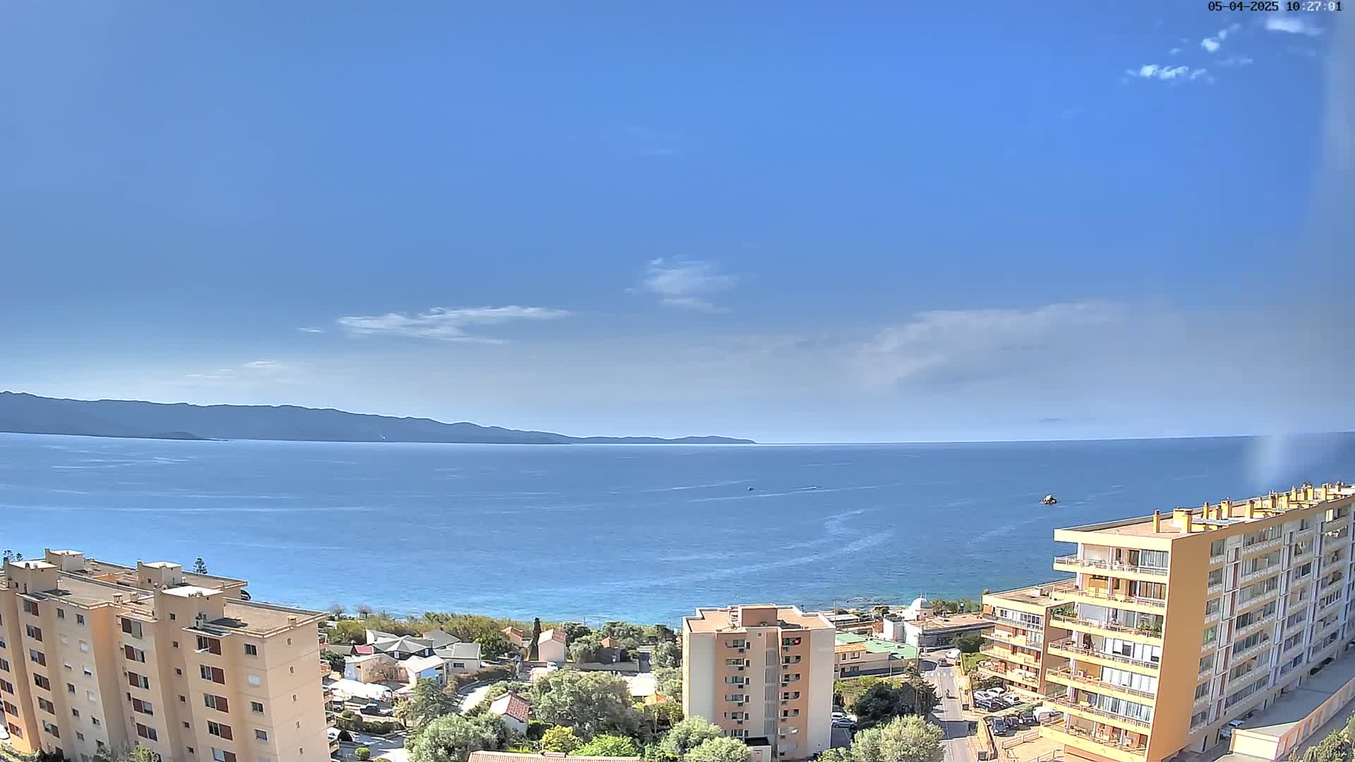 A clear, sunny day reveals a coastal scene with calm blue water, distant mountains, and several apartment buildings situated near a beach.