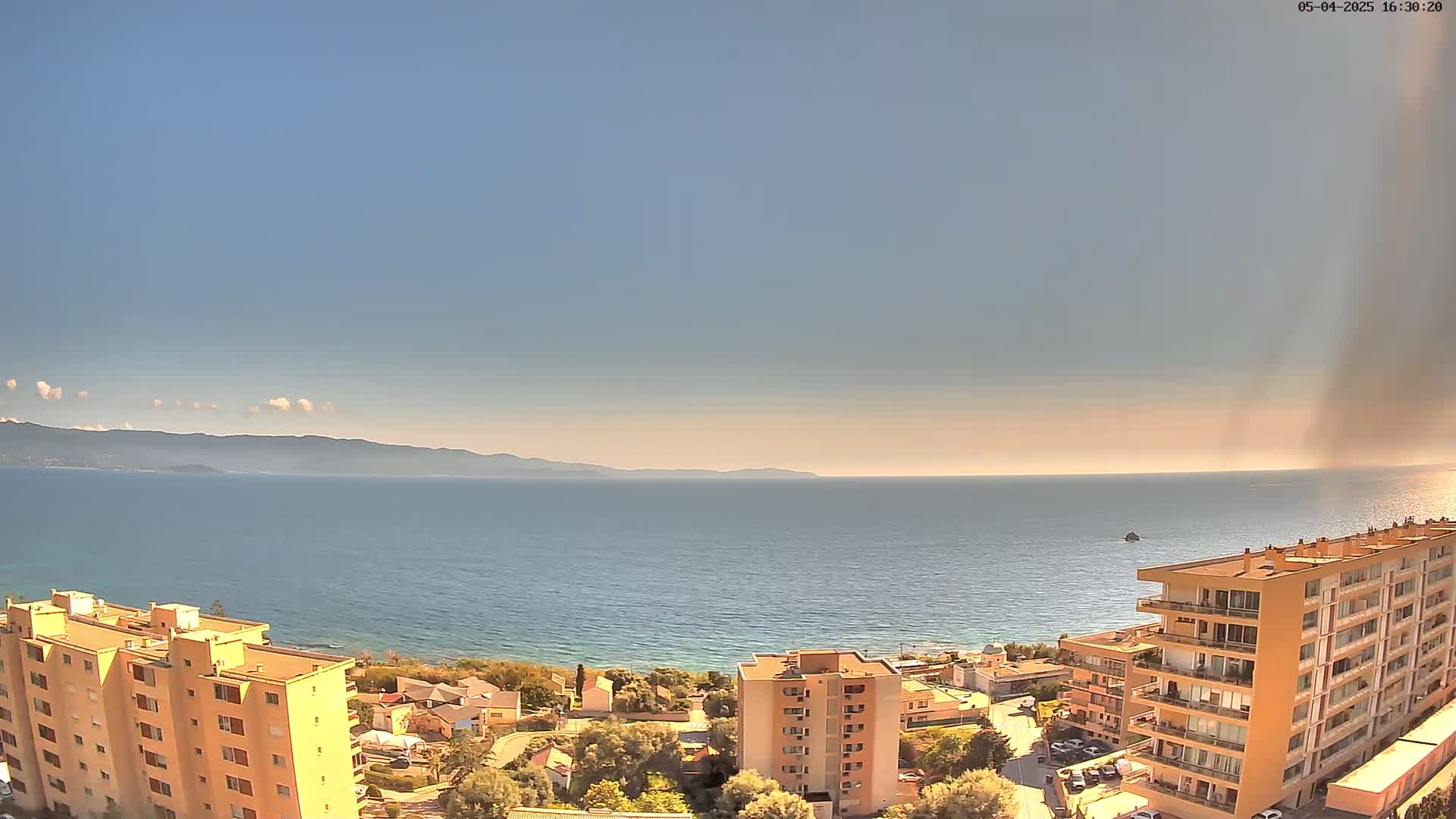 A coastal view shows a calm sea under a mostly clear sky, with several multi-story buildings and green vegetation in the foreground.
