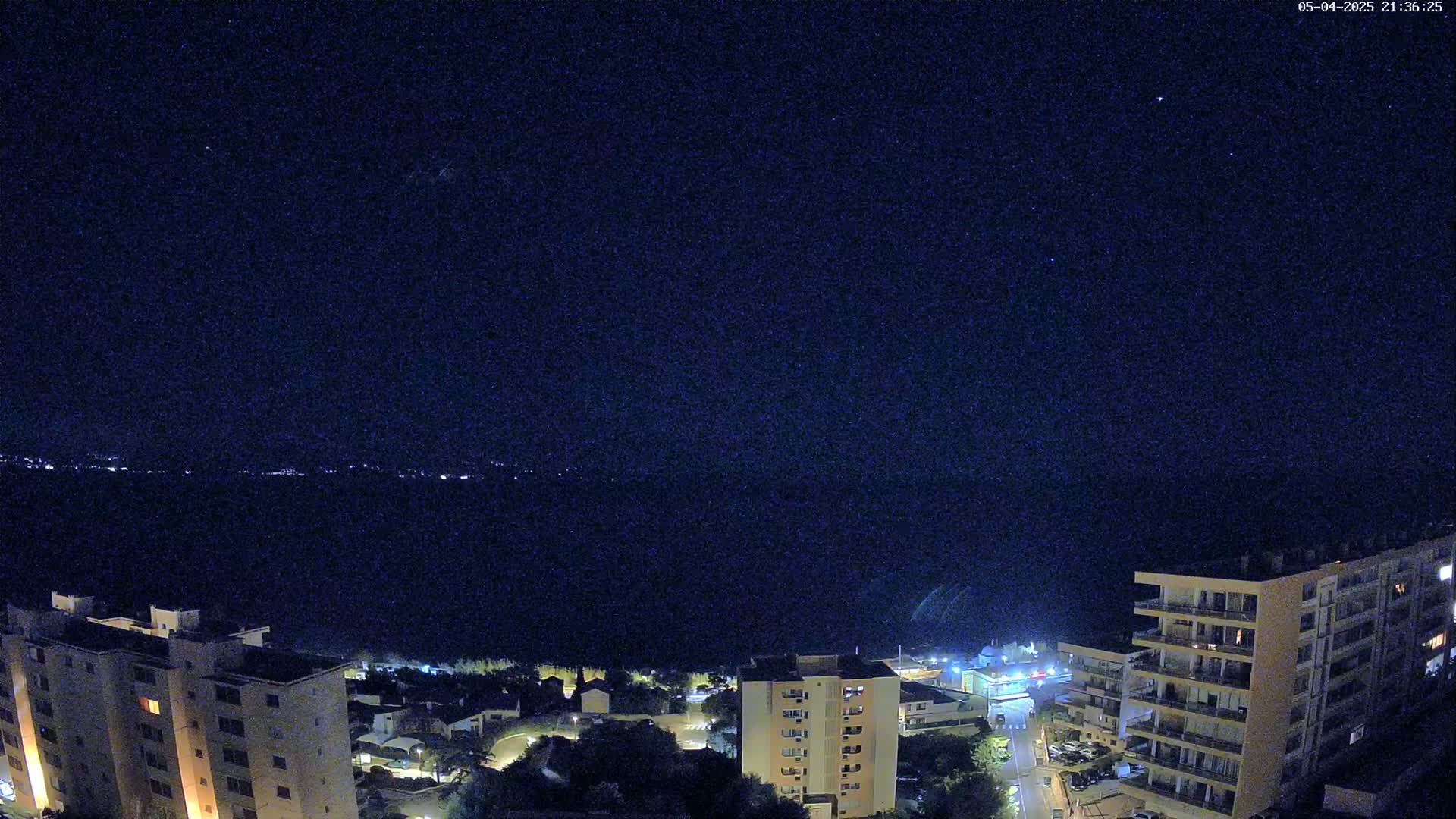 A nighttime, high-angle view shows several illuminated apartment buildings overlooking a dark body of water with a distant, faintly lit shoreline under a clear, starry sky.