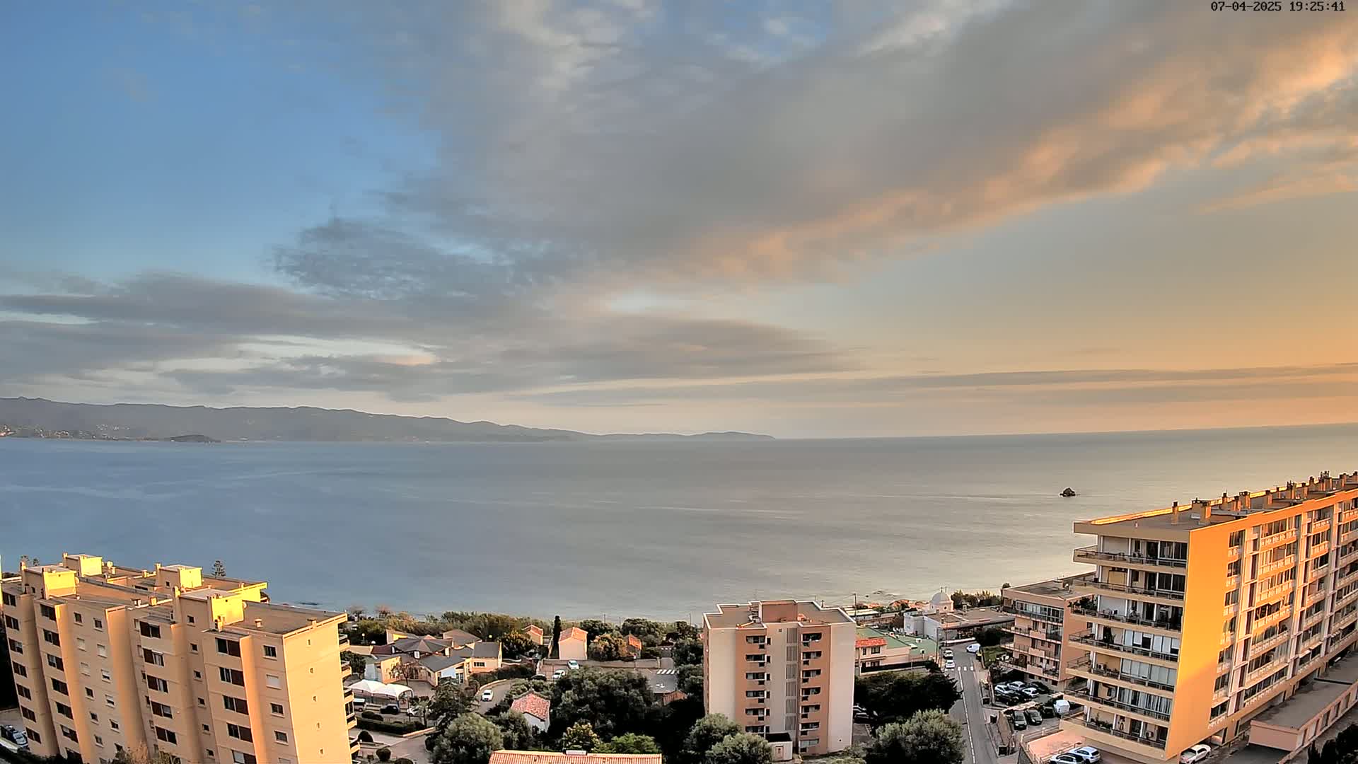 A calm sea stretches to distant mountains under a partly cloudy sunset sky, overlooking a coastal town with several multi-story buildings.