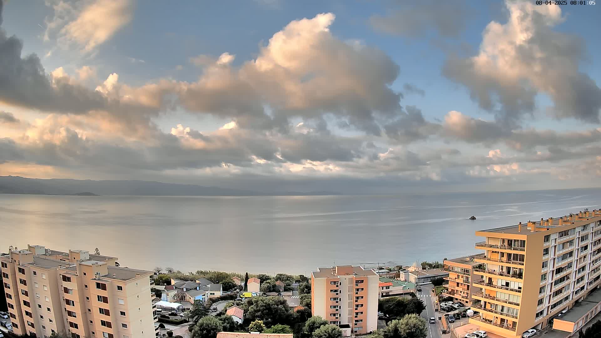 A partly cloudy sky overlooks a calm body of water and a coastal town with several apartment buildings.