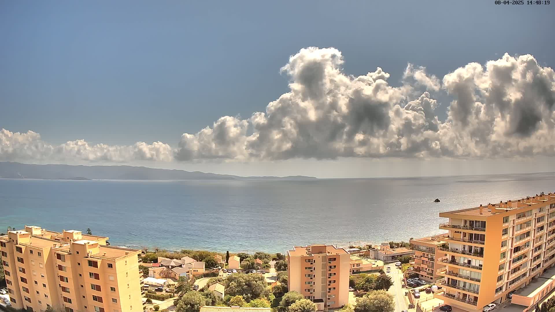 A mostly sunny day reveals a coastal view of apartment buildings, green vegetation, and a calm sea stretching to a distant mountain range under a partially cloudy sky.