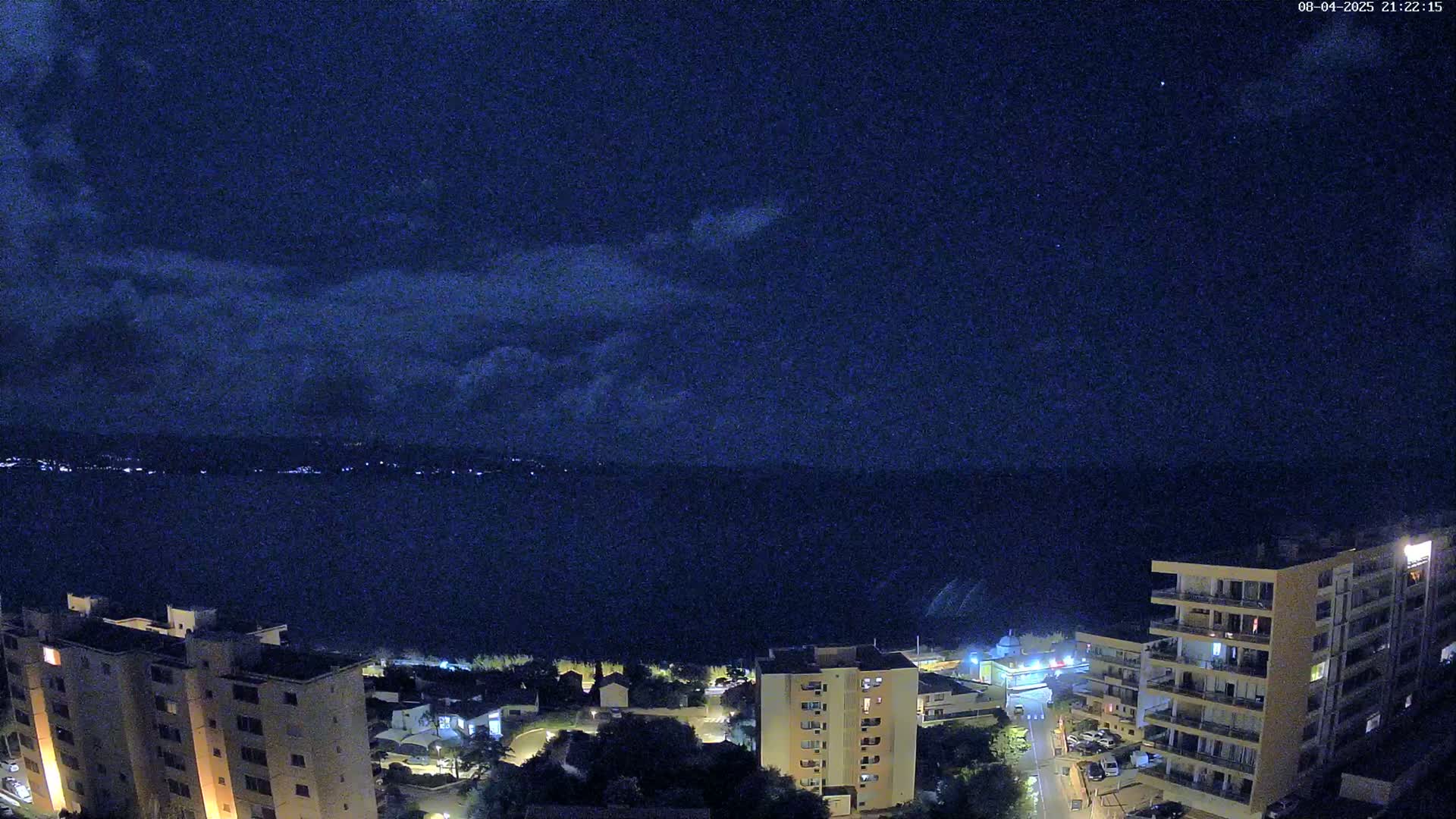 A nighttime view of a coastal town shows several apartment buildings overlooking a dark body of water under a partly cloudy sky.