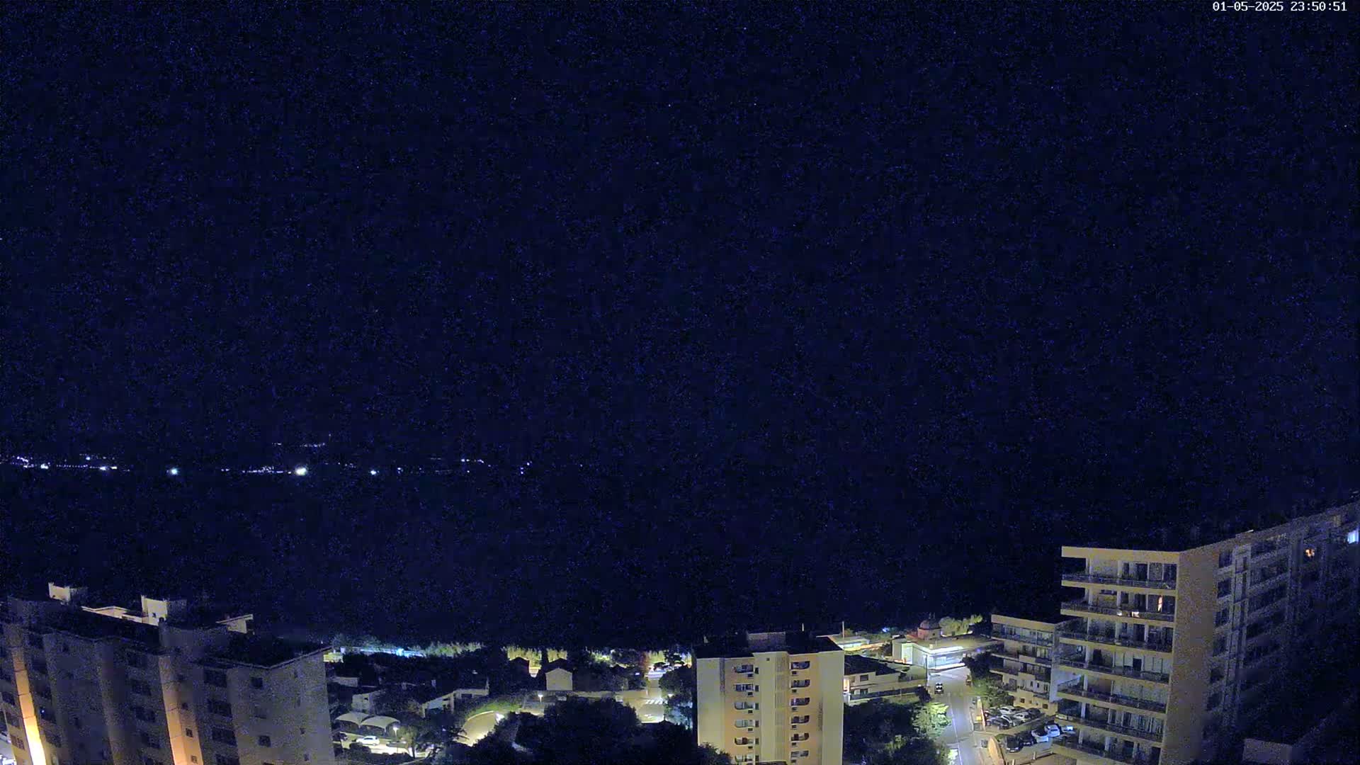 A nighttime view of a city skyline with apartment buildings and a dark body of water in the distance under a clear, starry sky.