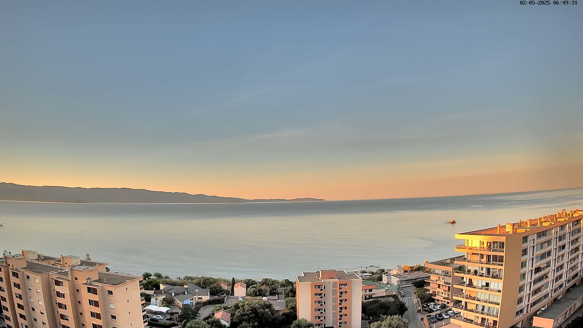 A calm sea at sunset is viewed from above a coastal town, with mountains visible in the distance.