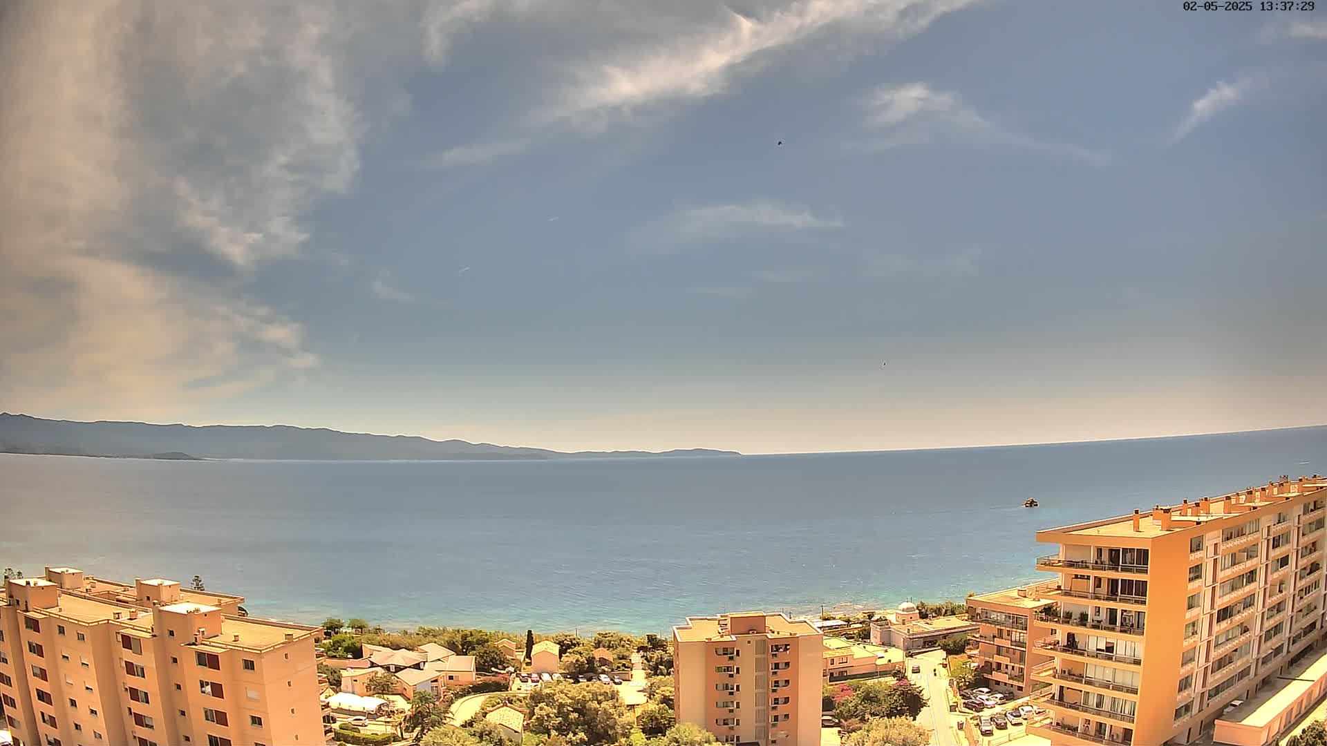 An elevated view shows a calm, blue sea extending to a distant mountain range under a partly cloudy sky, with several apartment buildings situated on the coastline.
