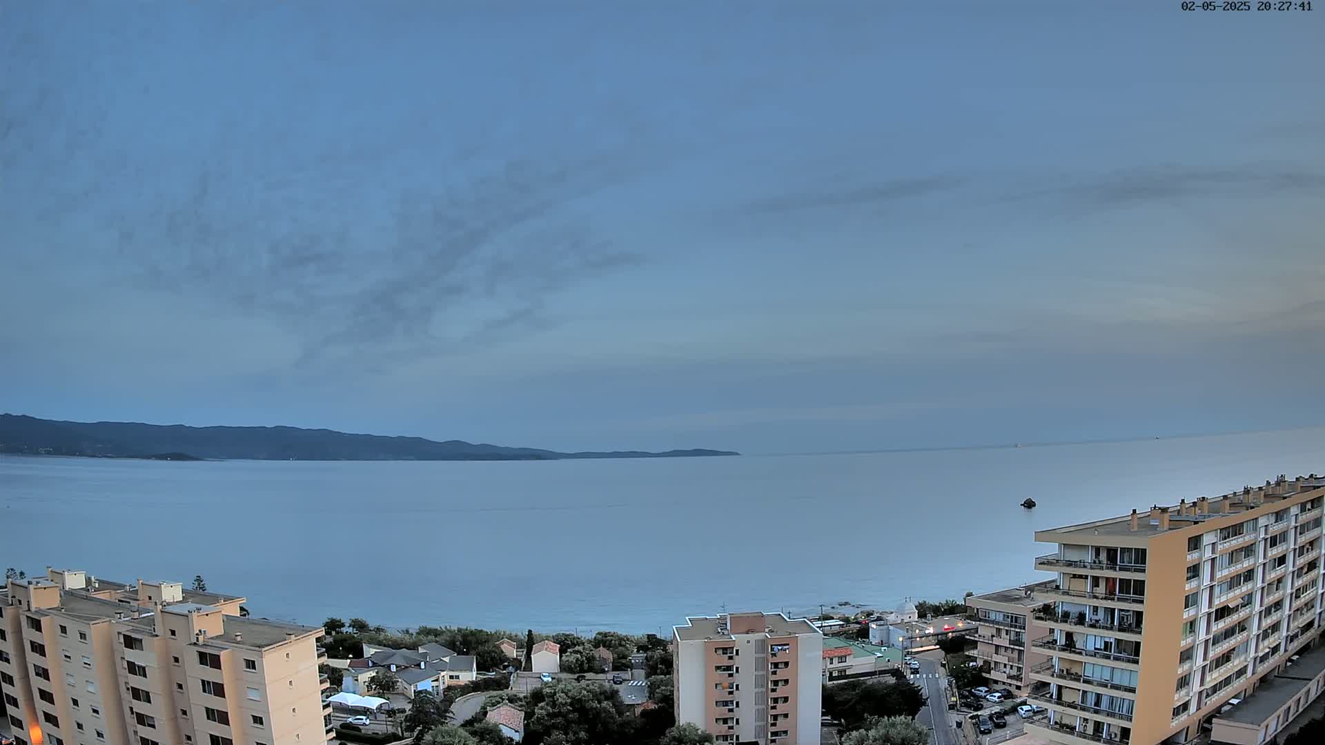 A calm sea stretches out beneath a mostly clear, twilight sky, overlooking a coastal town with several apartment buildings.