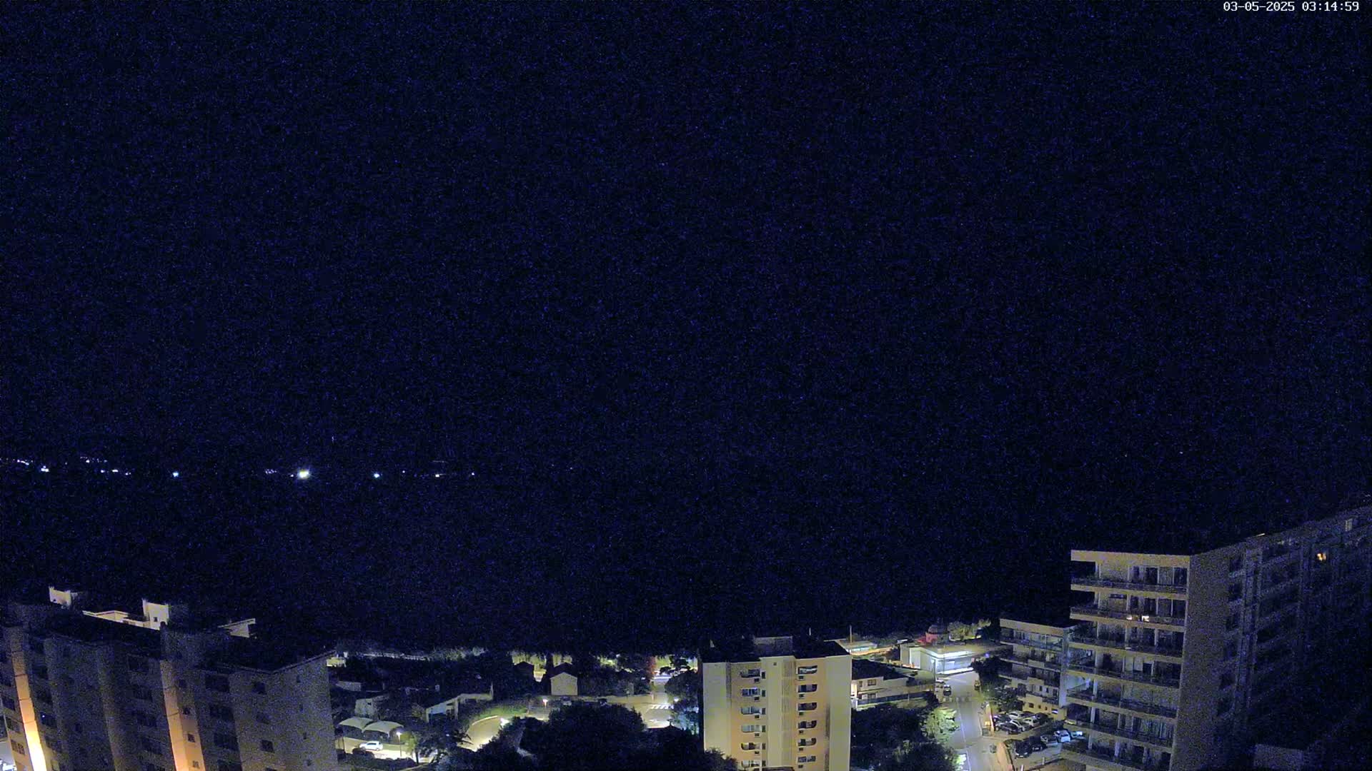 A nighttime, high-angle view shows several apartment buildings illuminated near a dark body of water with faint lights visible on the opposite shore.