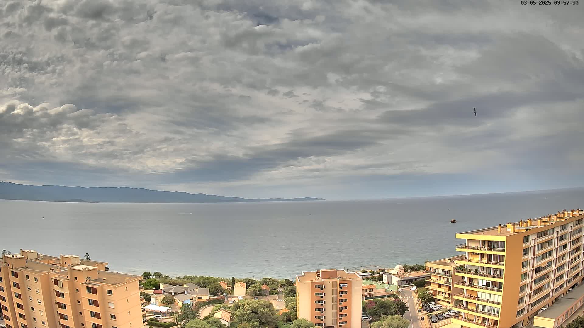 A coastal scene under a cloudy sky shows a calm sea, distant mountains, and several apartment buildings near the shore.