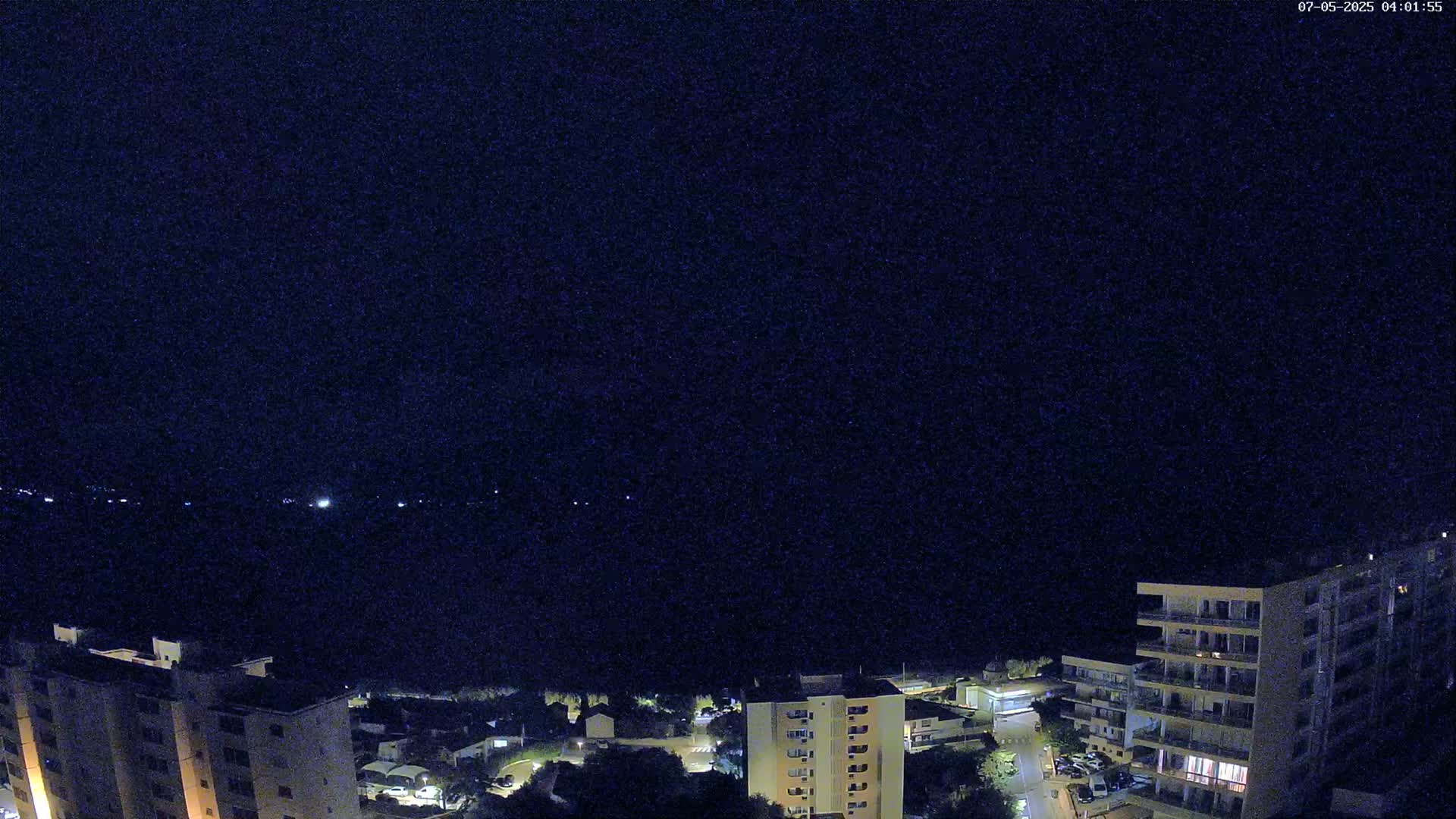 A nighttime, high-angle view shows several apartment buildings illuminated against a dark, clear sky, with a body of water visible in the distance.