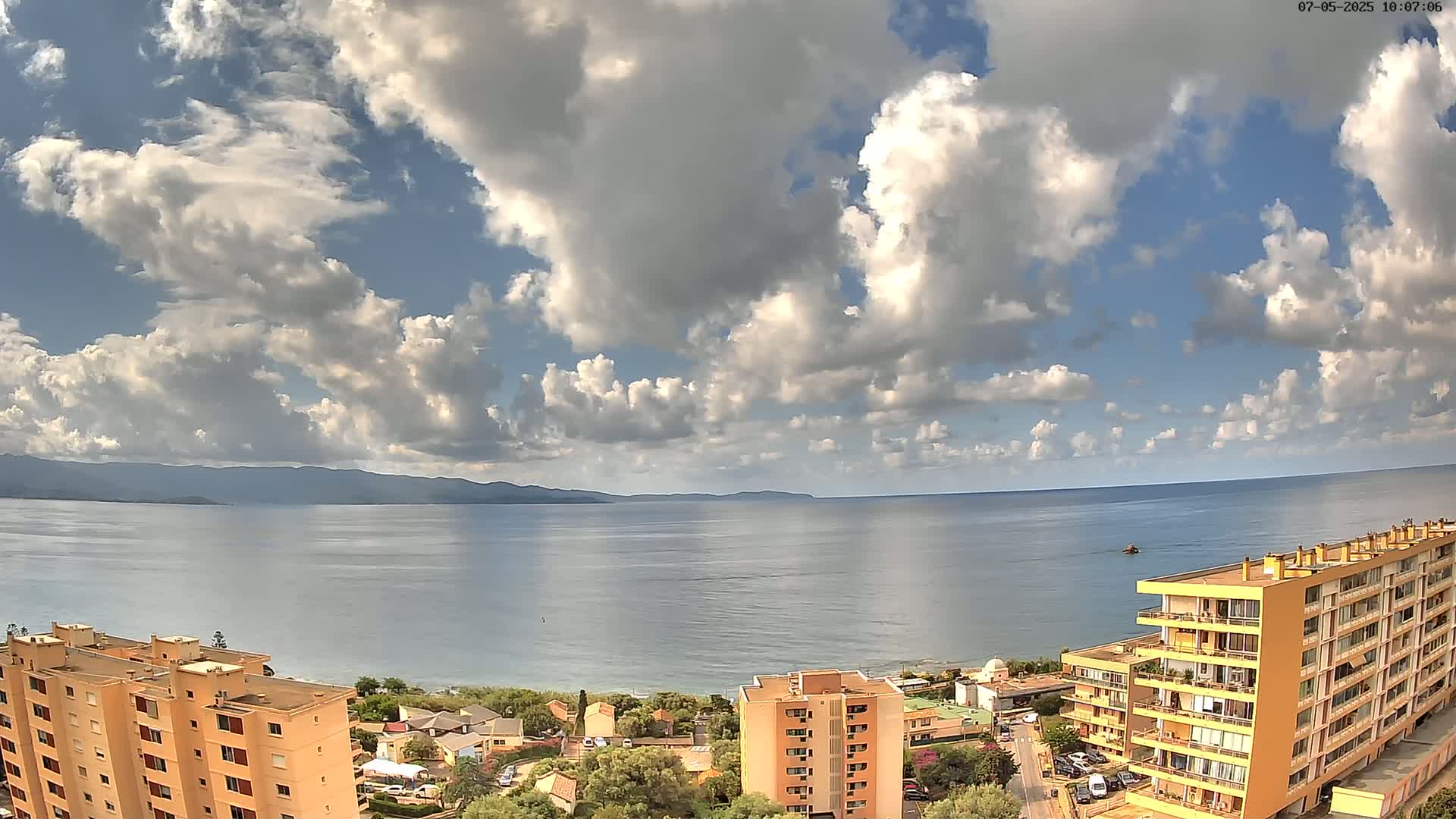 A partly cloudy sky overlooks a calm sea with a distant mountain range and coastal buildings.