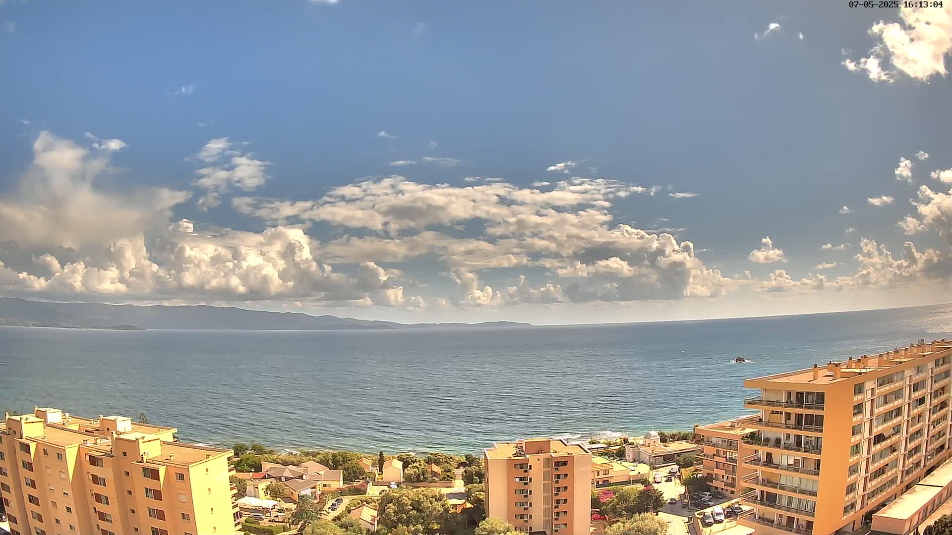 A partly cloudy sky overlooks a calm sea and a coastal town with several multi-story buildings.
