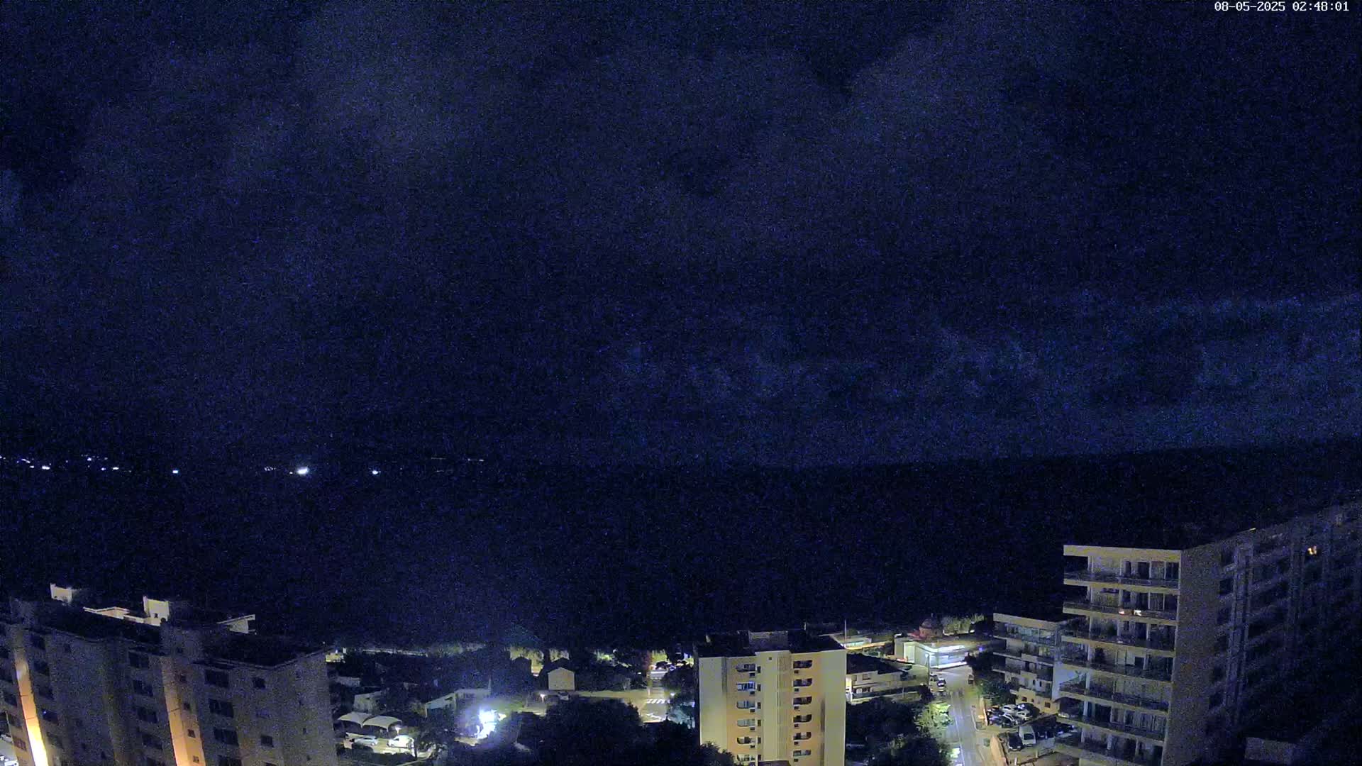 A nighttime view of a coastline with apartment buildings, showing a dark, cloudy sky over a dark body of water.