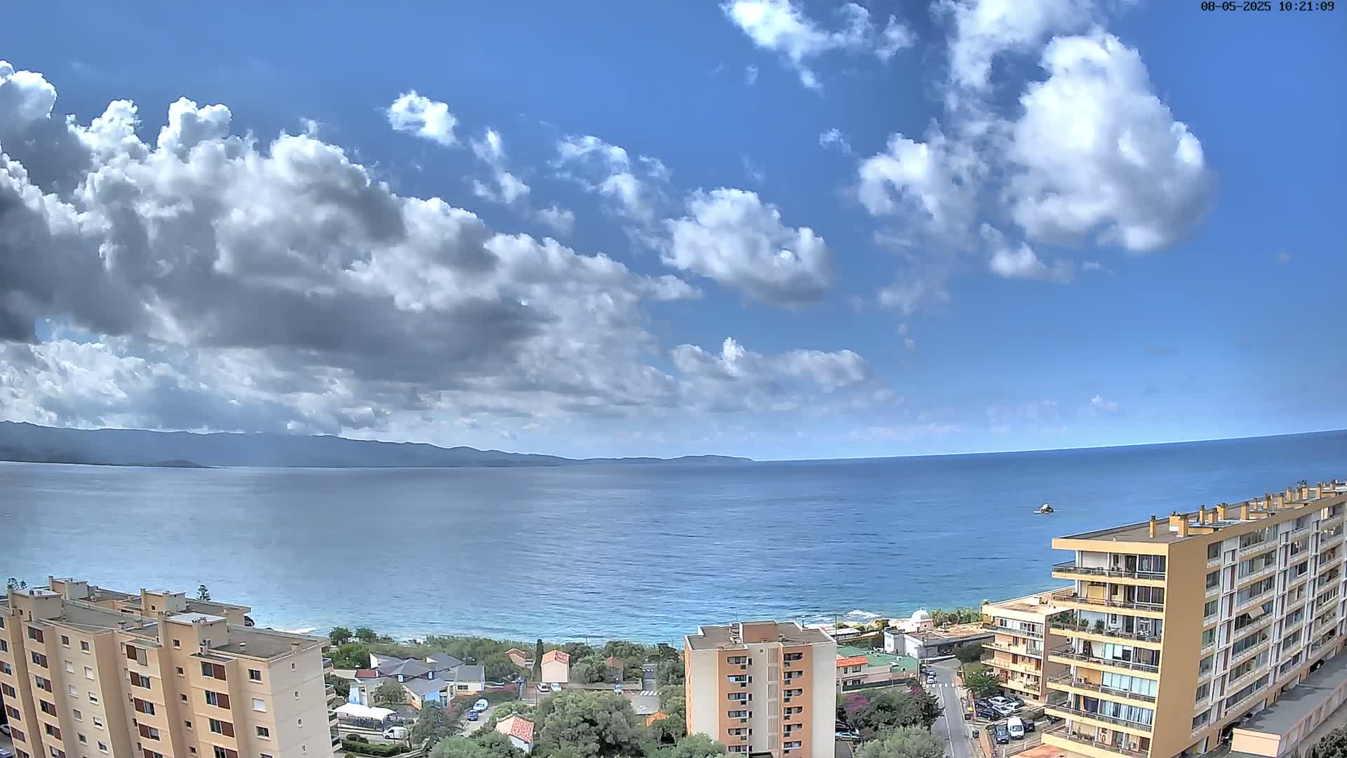 A partly cloudy blue sky overlooks a calm ocean, a shoreline with buildings, and distant mountains.