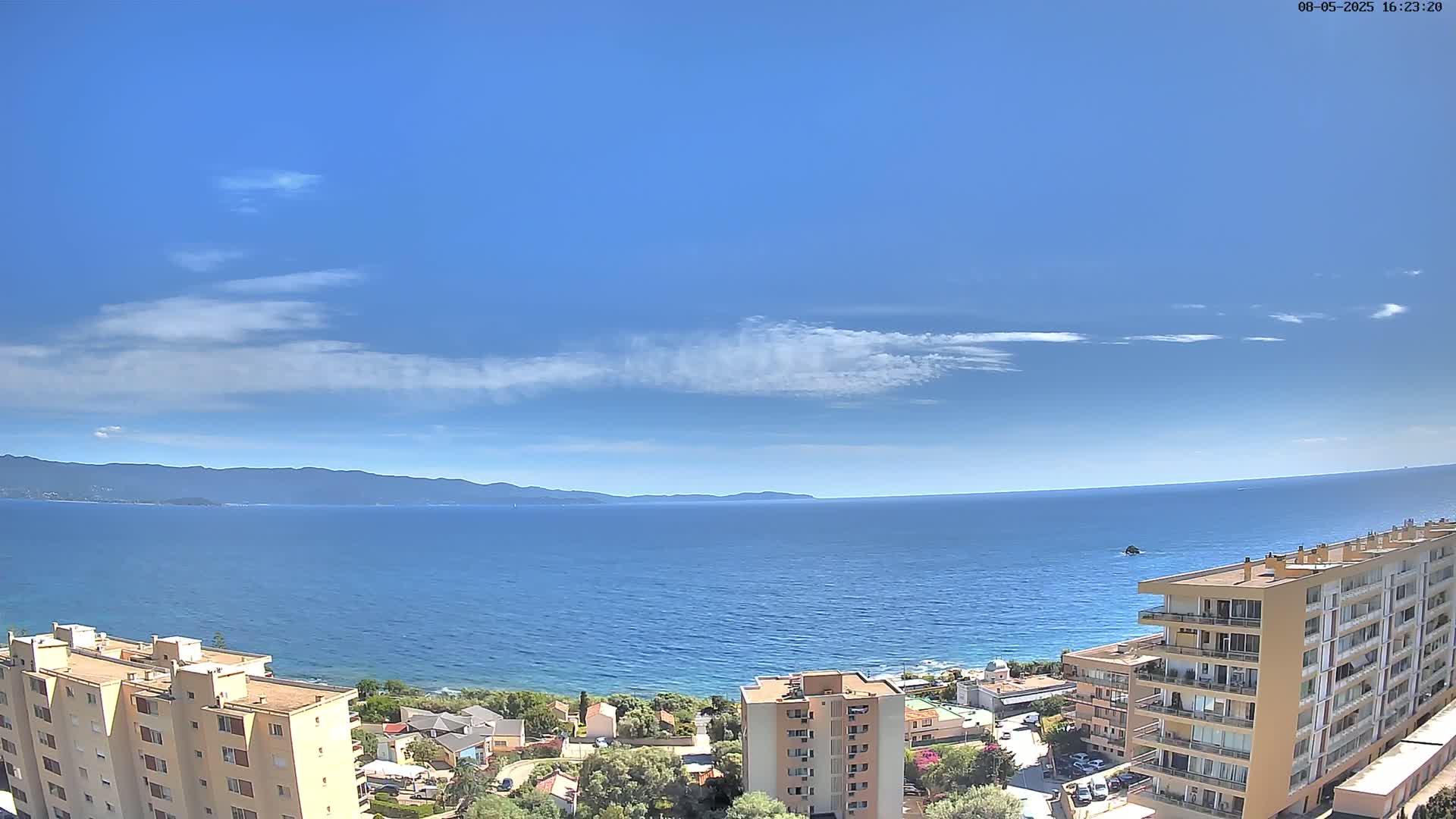 A mostly sunny day shows a calm blue sea extending to distant mountains under a mostly clear sky, with several apartment buildings in the foreground.