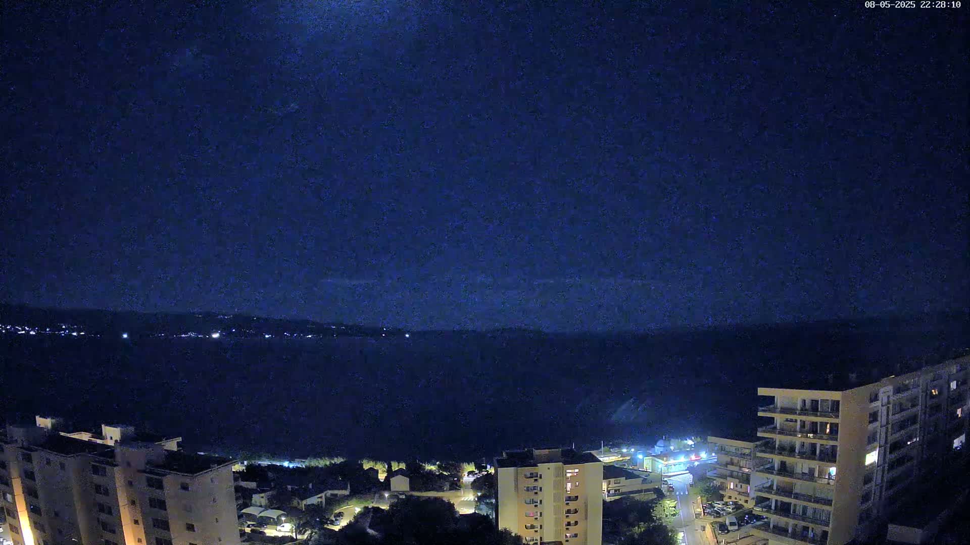 A nighttime view shows a dark body of water extending to a distant, dimly lit shoreline under a clear, dark blue sky.